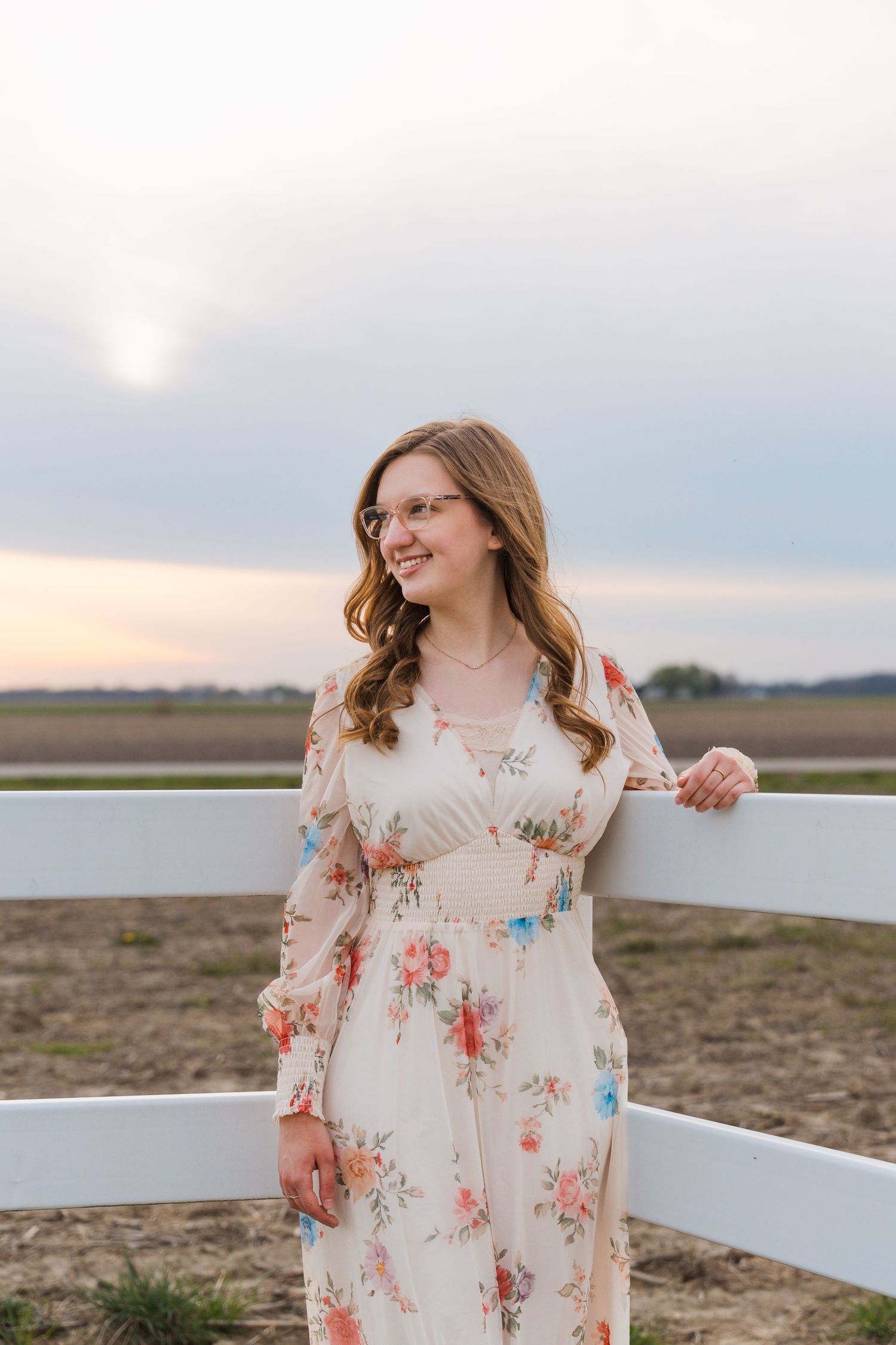 Young woman in floral maxi dress leaning on white fence at sunset in a rural field.