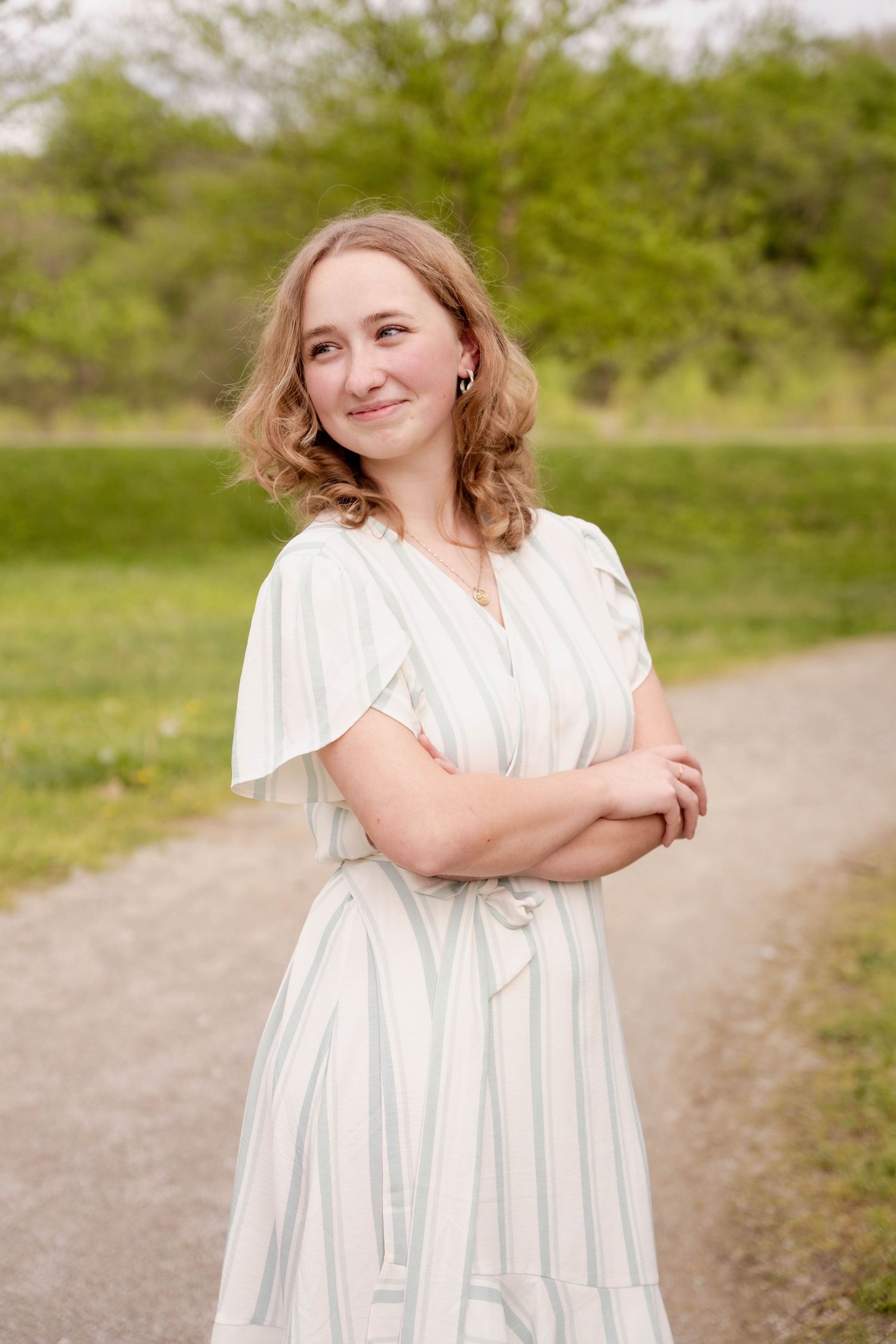 A person in a white summer dress stands on a gravel path with arms crossed and greenery in the background.