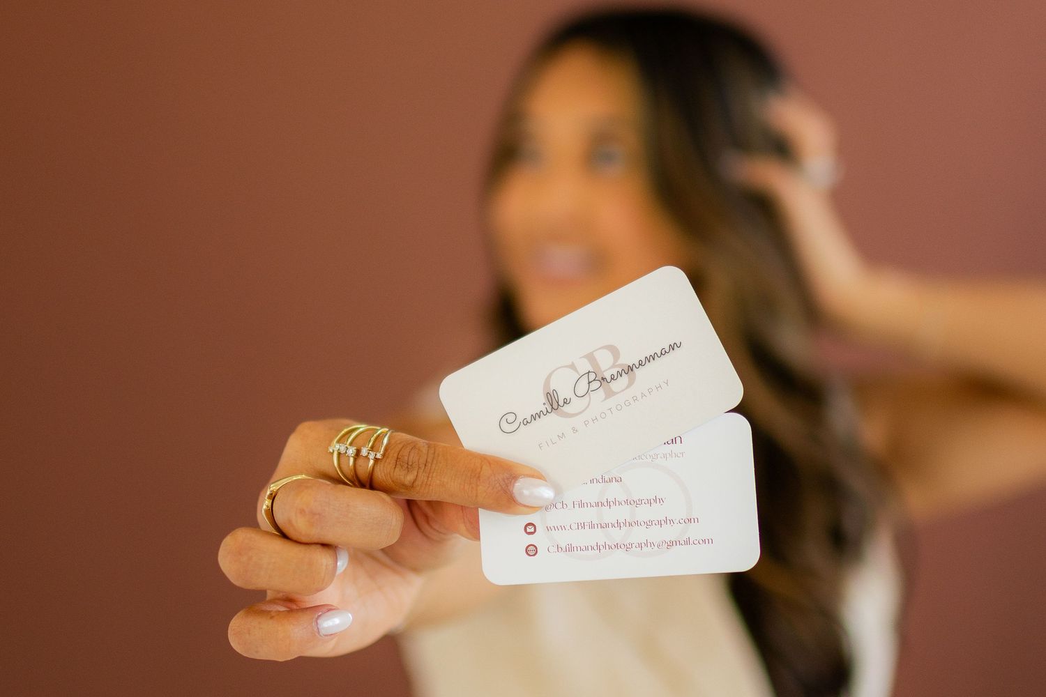 Smiling woman with long brown hair holds out two white business cards toward the camera, wearing gold rings against a terracotta wall.