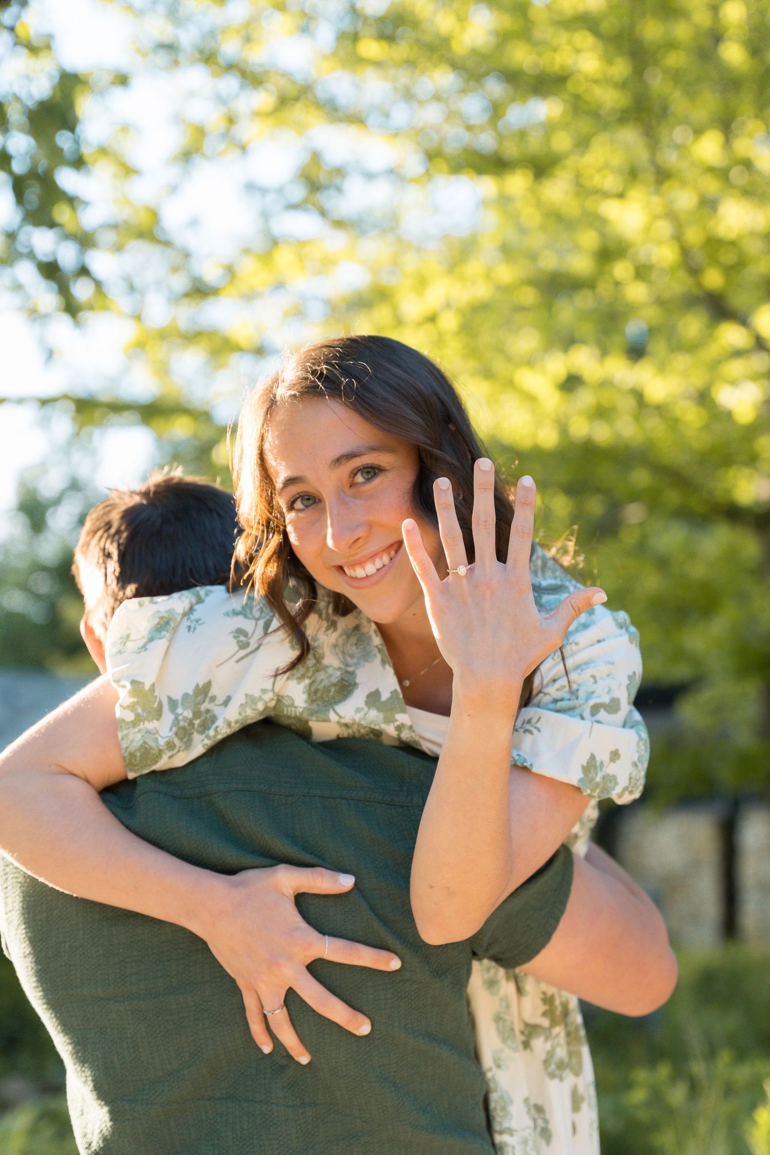 Smiling woman hugging a man outdoors while showing off her engagement ring, with green trees in the background.