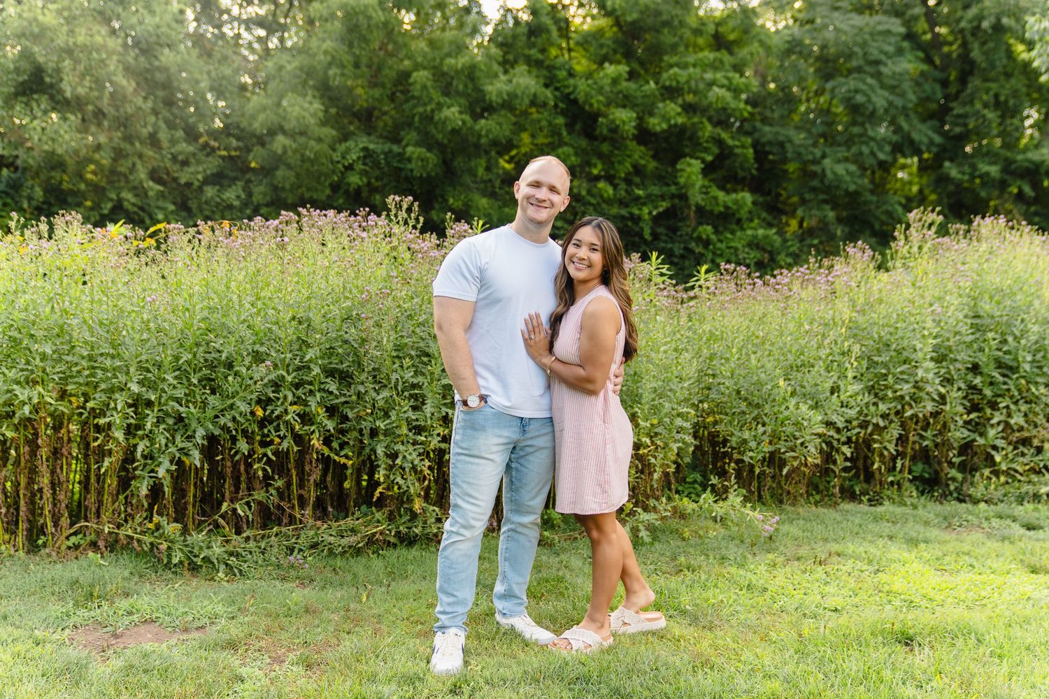 Couple smiling together outdoors in a lush green field, man in white shirt and jeans, woman in pink dress.