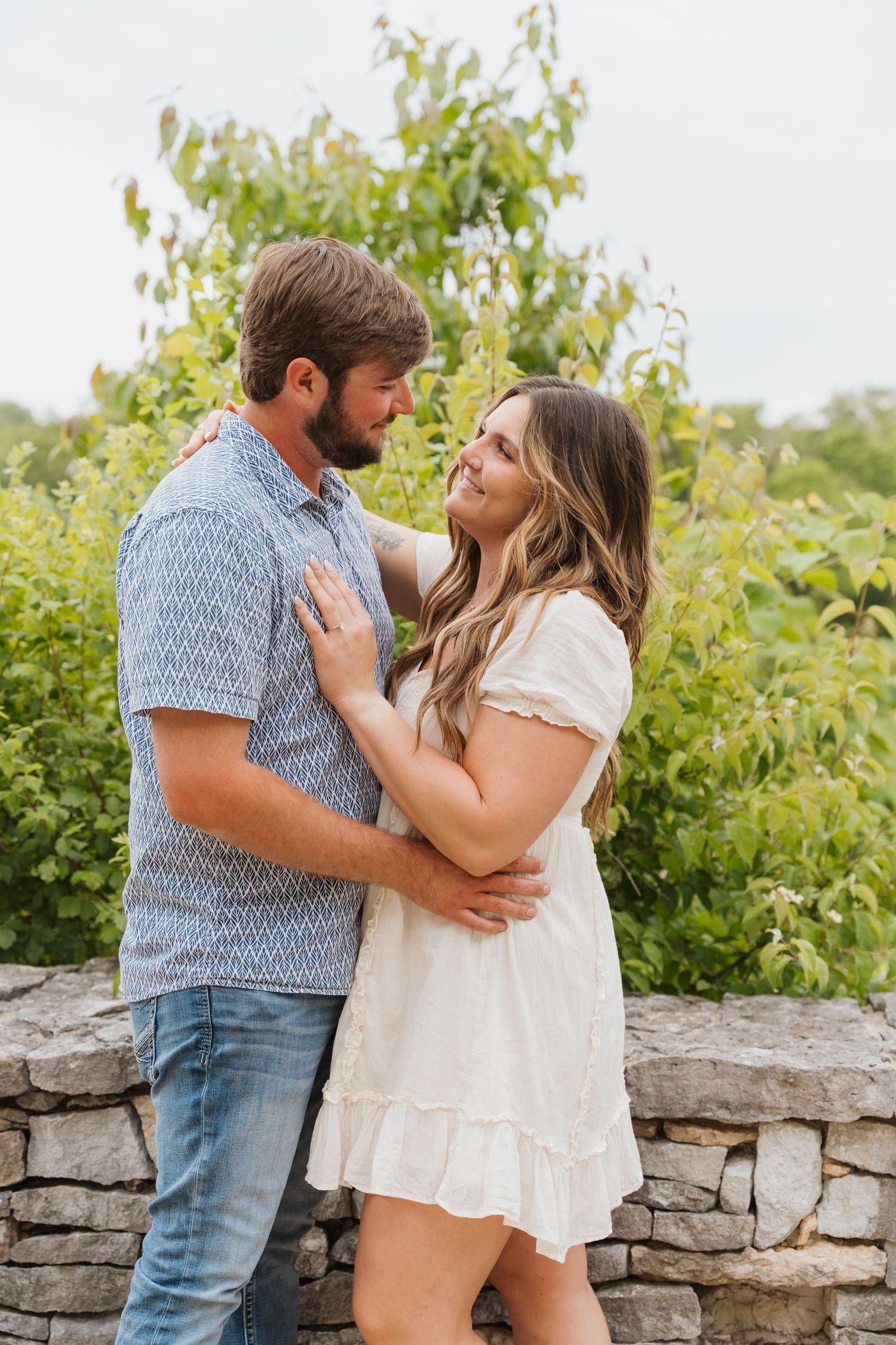 Couple embracing outdoors near a stone wall, man in blue shirt and woman in white dress, surrounded by lush greenery.