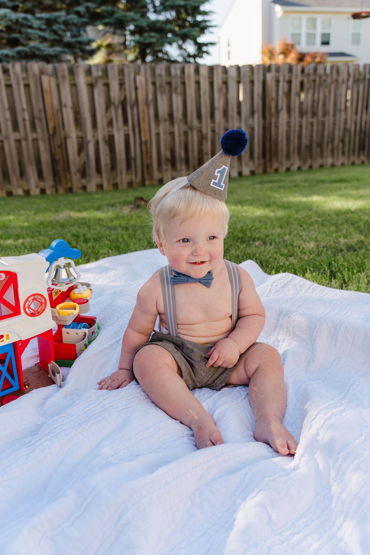 Smiling baby boy in suspenders and a number one party hat sits on a white blanket outdoors for his first birthday.