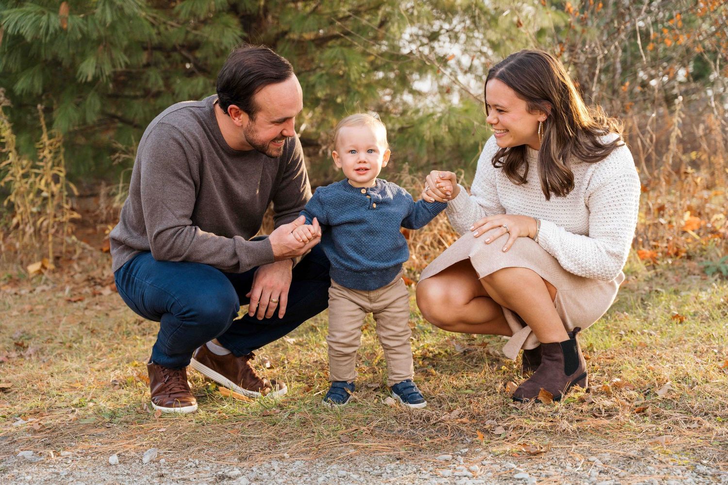 Smiling couple crouching outdoors in an autumn field holding the hands of a toddler boy standing between them during a family photo session.