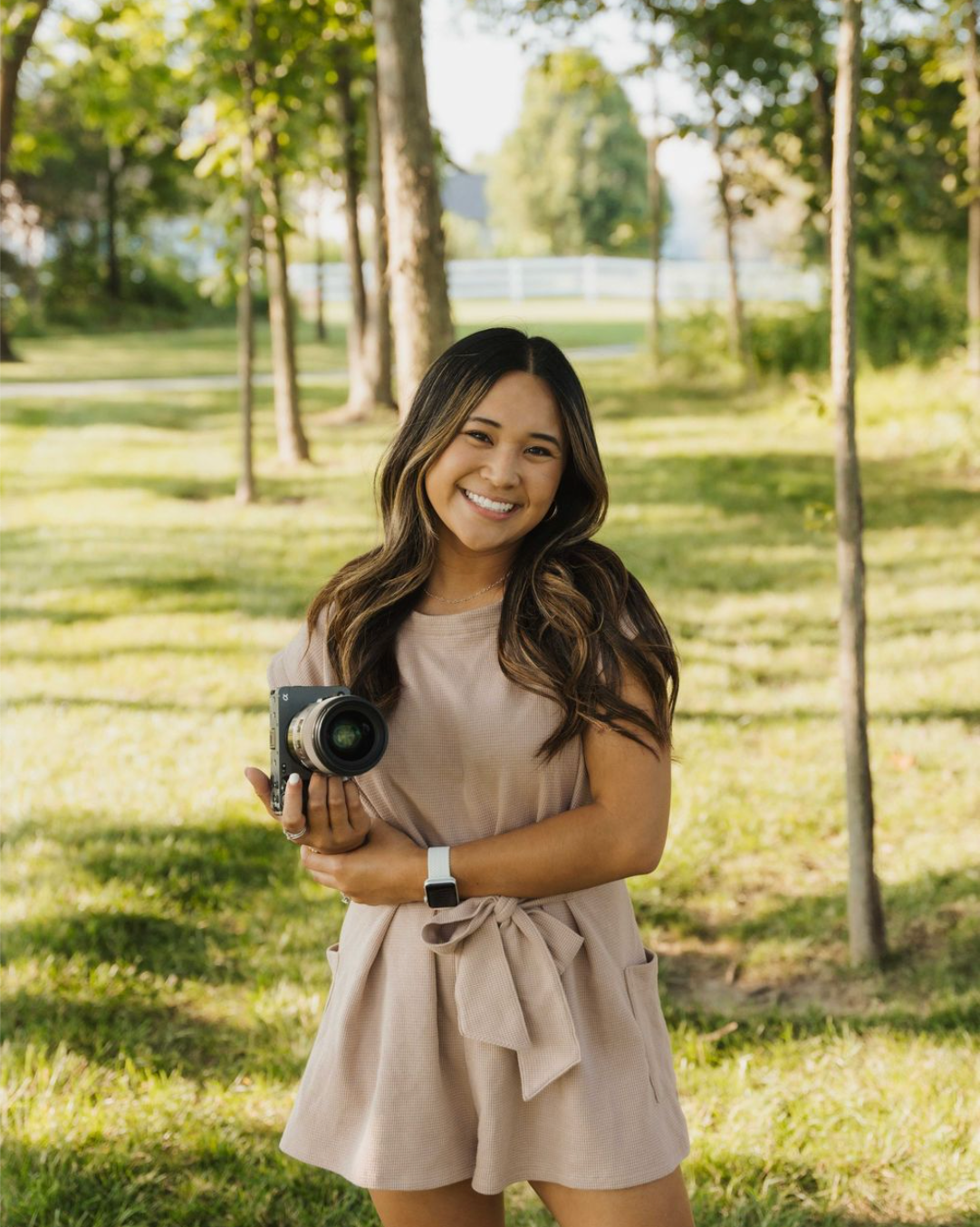 Woman in beige ruffled dress poses outdoors in a sunny park setting with trees in the background.