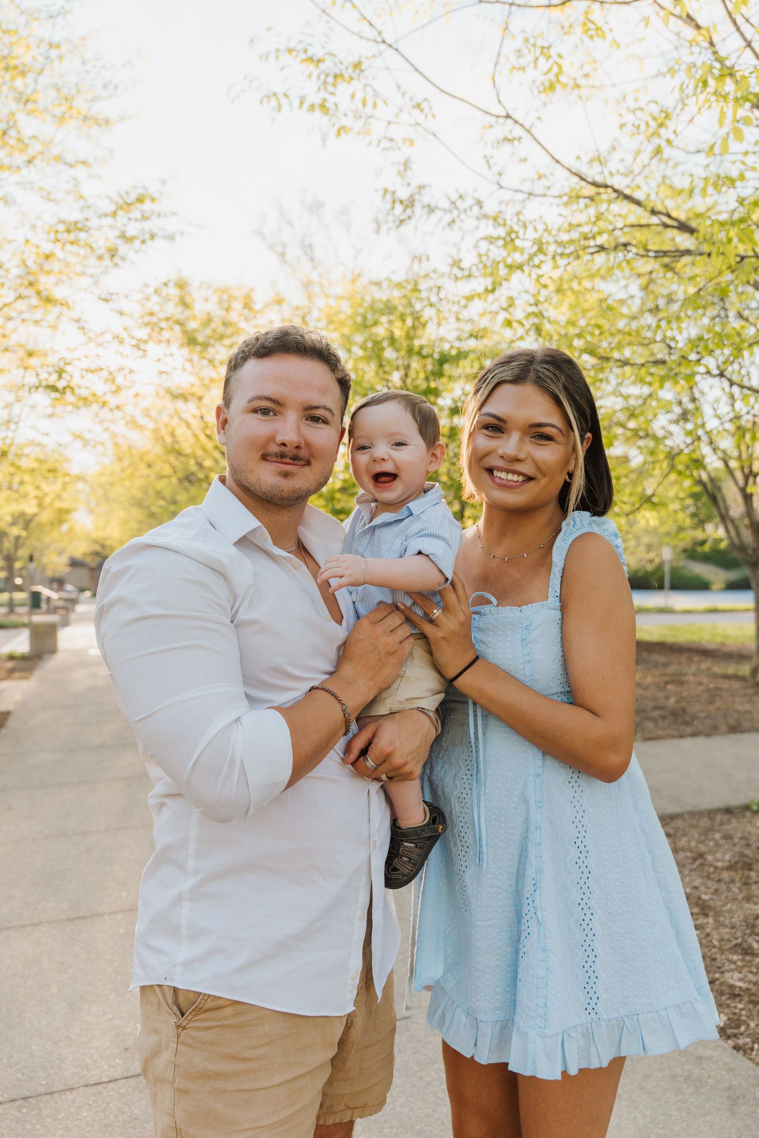 Happy young family with parents holding smiling baby boy outdoors in a sunny park setting.