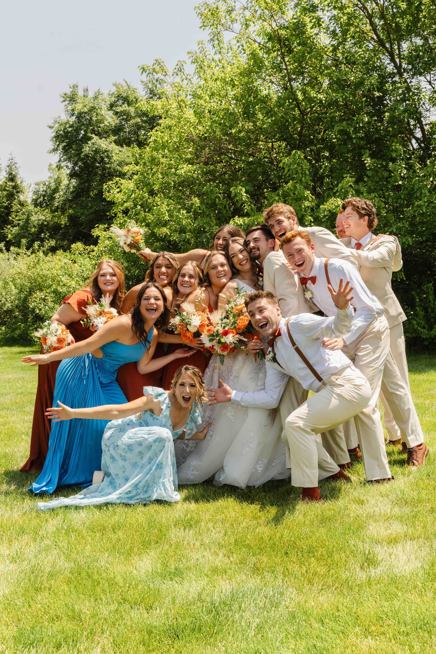Joyful wedding party posing outdoors on green lawn, bride in white gown with orange bouquet surrounded by bridesmaids and groomsmen.