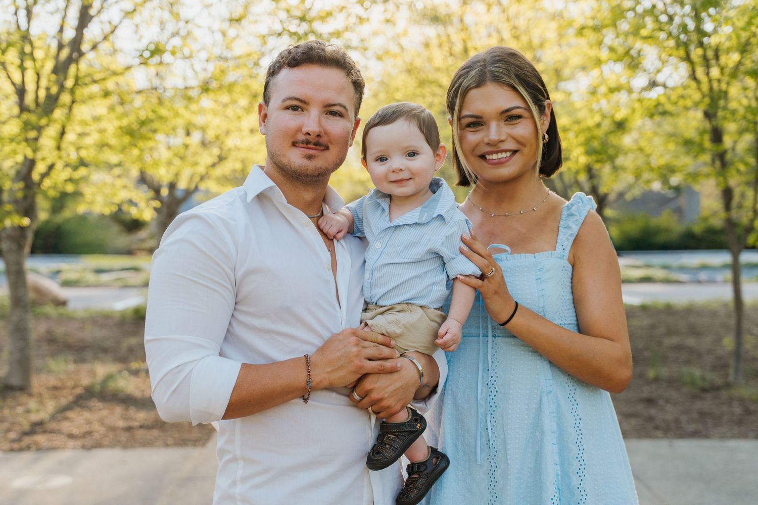 A smiling couple holds their baby boy outdoors, dressed in casual attire with lush green trees in the background.