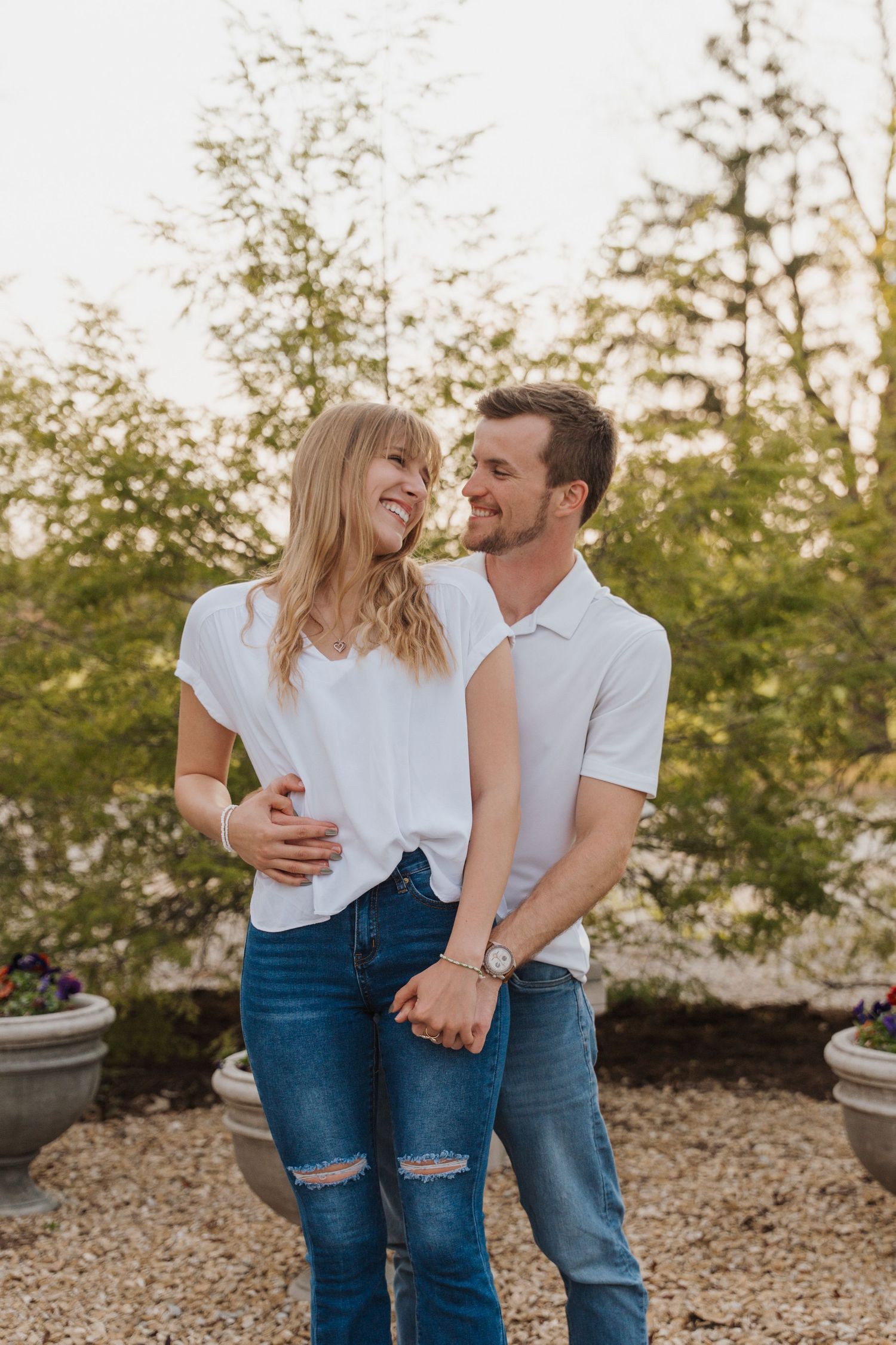 A smiling couple in matching white tops and jeans embrace outdoors in a garden setting.