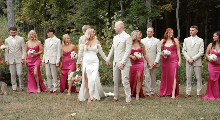 Wedding party in pink and beige attire poses together in a line on grassy lawn during outdoor celebration.