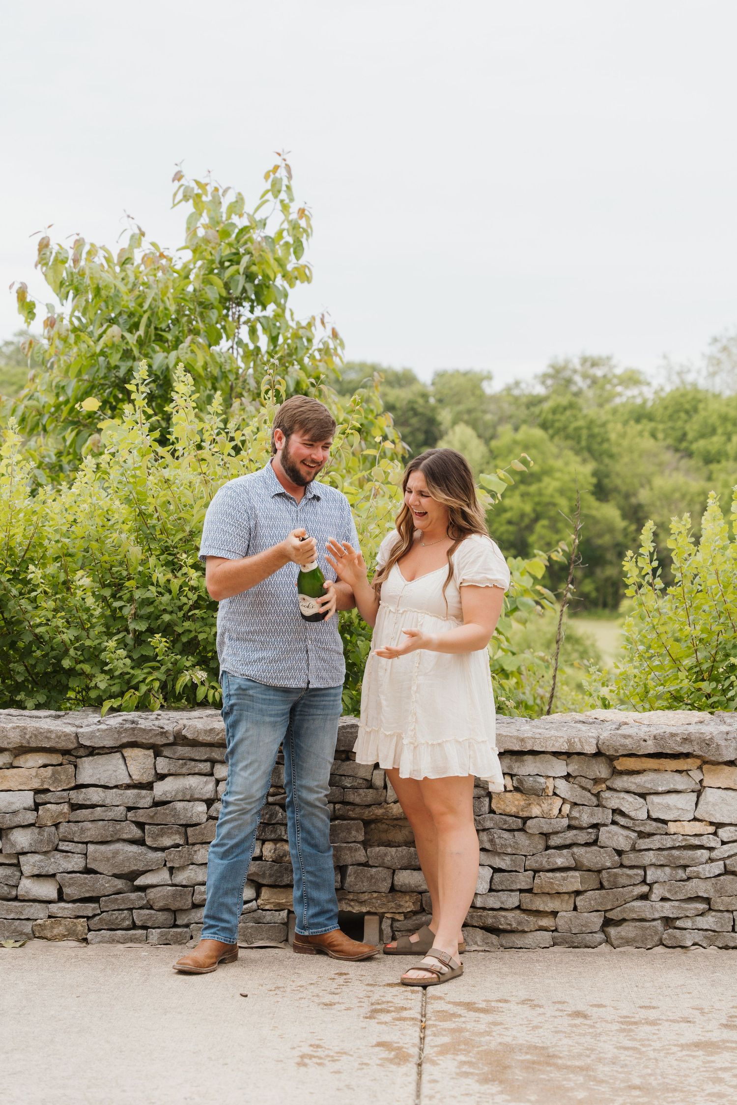 Couple celebrating engagement by opening champagne bottle outdoors near stone wall with lush green trees.