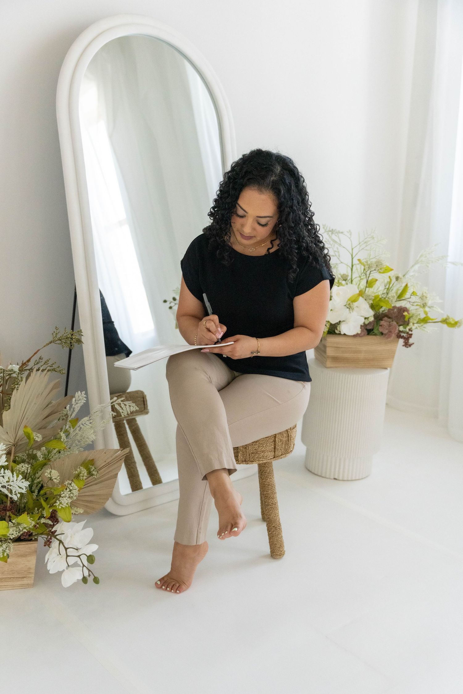 A person in a black top and beige pants sits on a wooden stool in a bright white studio with floral decor.