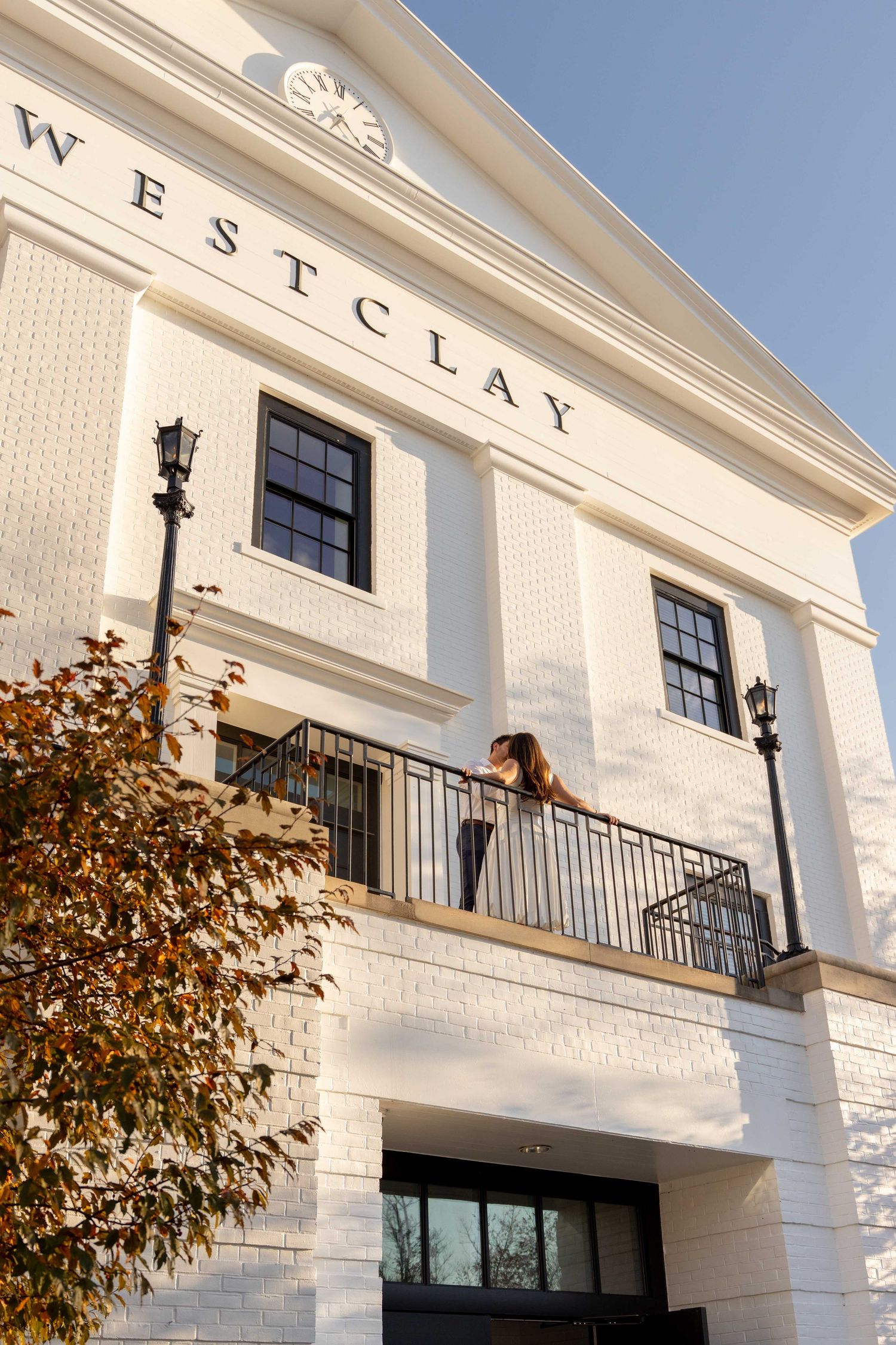 White brick building facade with black windows, balconies and decorative lighting fixtures against a blue sky.