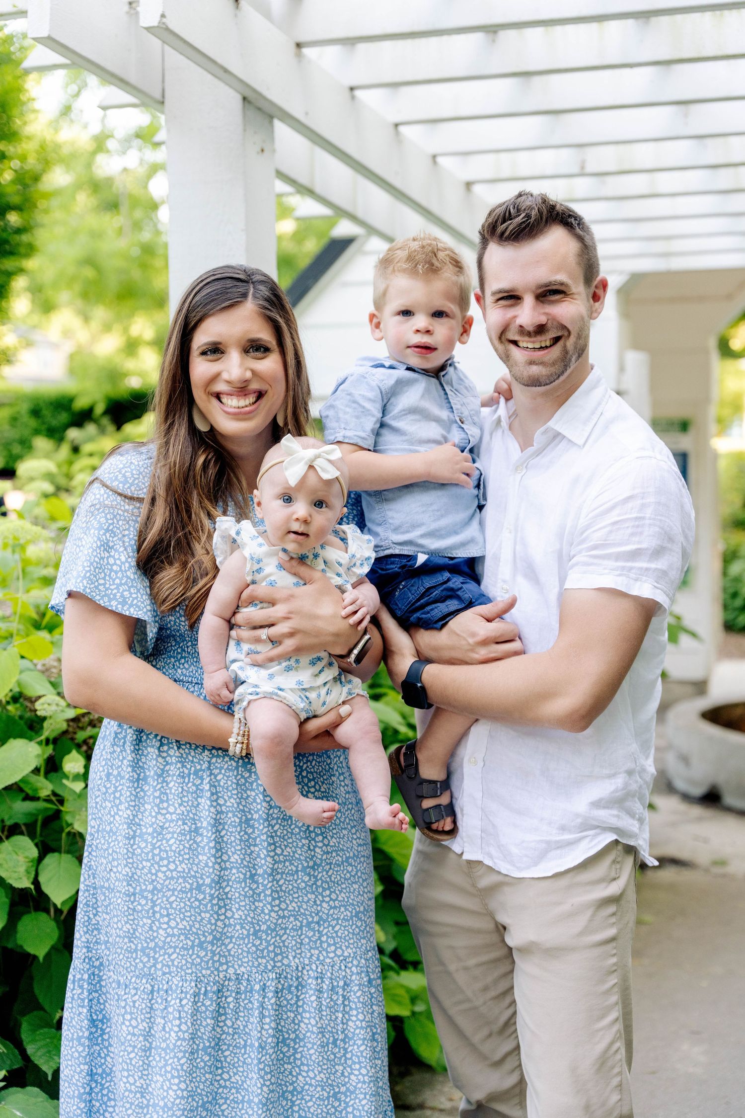 A smiling family of four poses outdoors, with parents holding a baby girl and toddler boy in a lush garden setting.