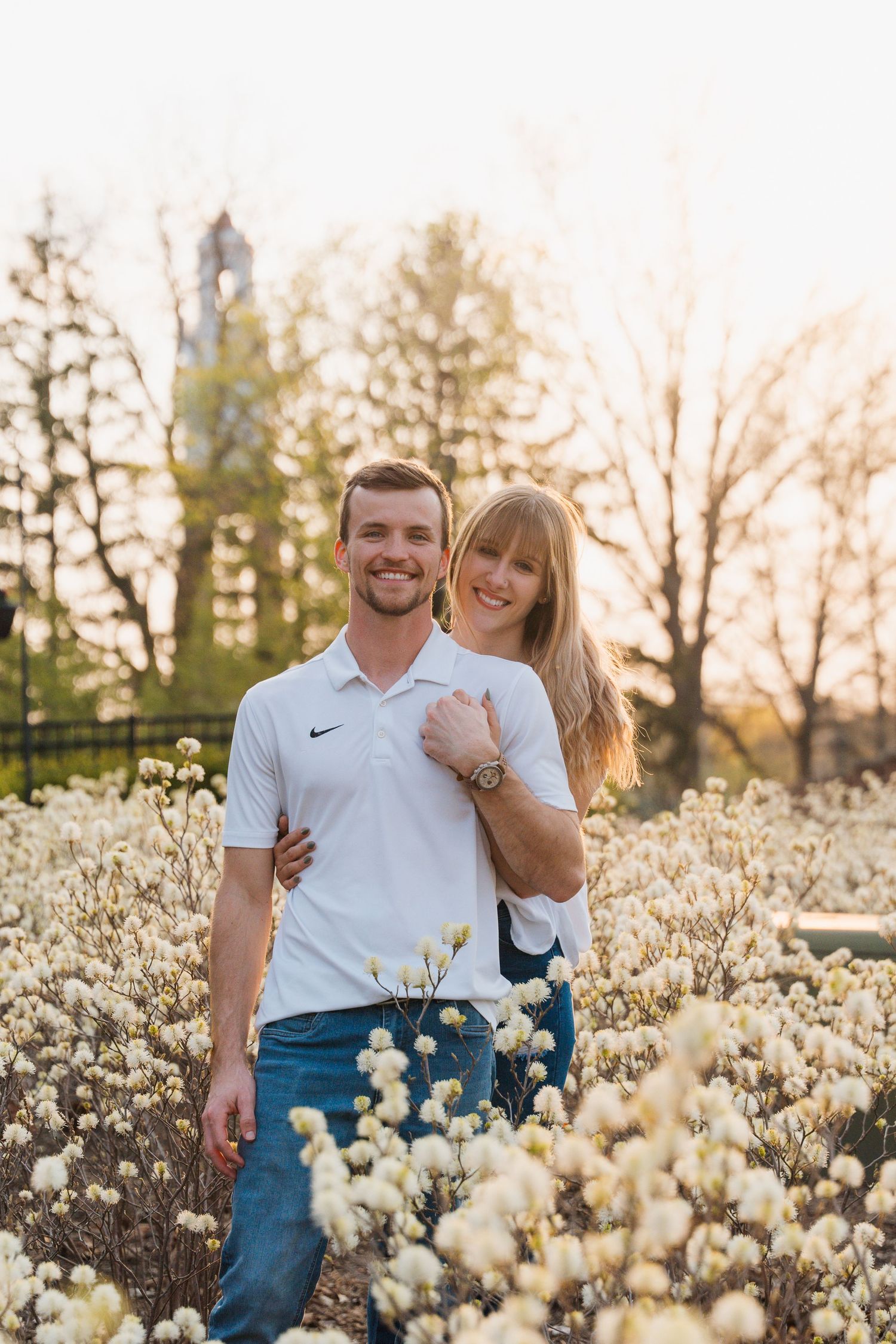 Couple smiling together in a field of white wildflowers at golden hour with trees and a tower in the background.