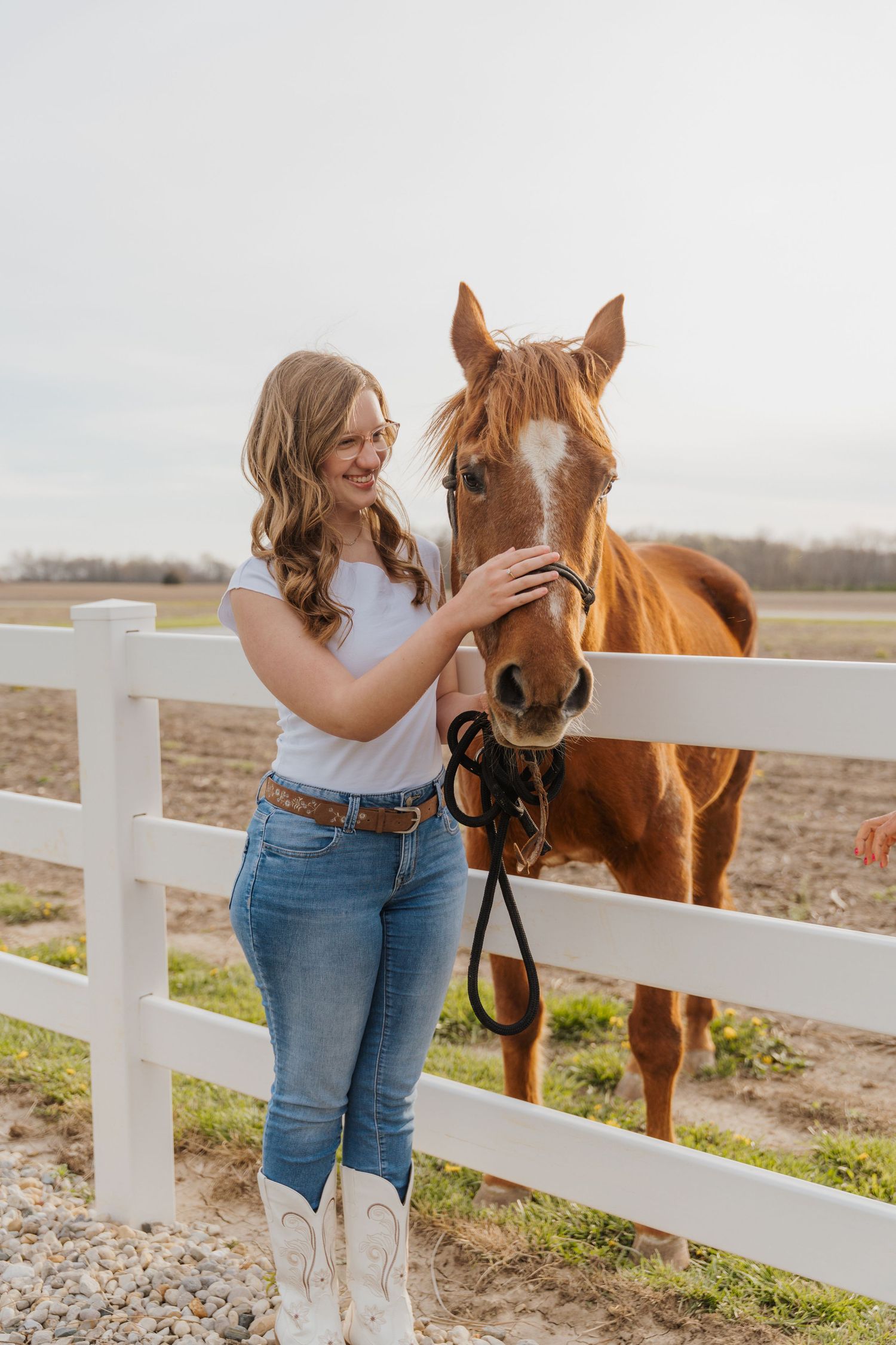 Young woman in white tee and jeans petting a chestnut horse by a white fence on a farm.