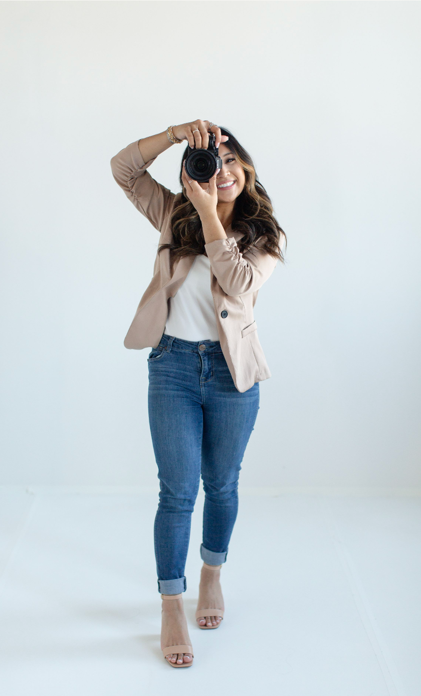 Photographer in beige blazer, white top and rolled jeans takes photos while kneeling on white backdrop.