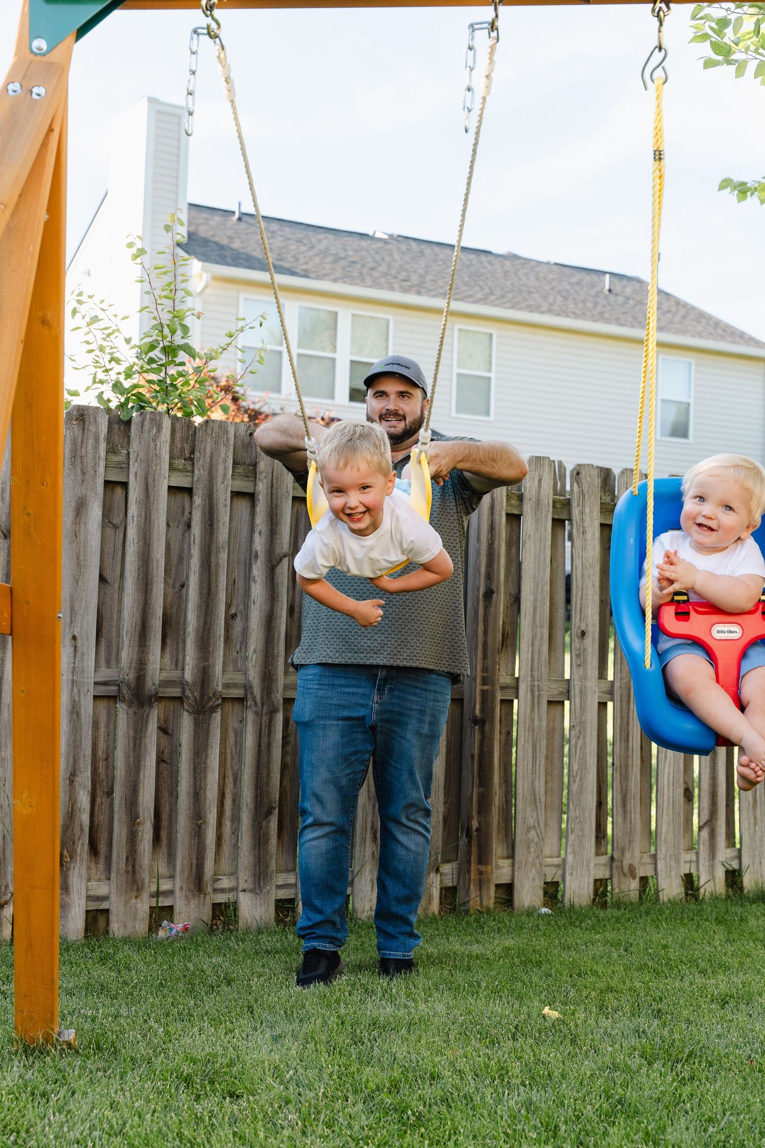 A father joyfully pushes two young children on backyard swings, all smiling on a sunny day.