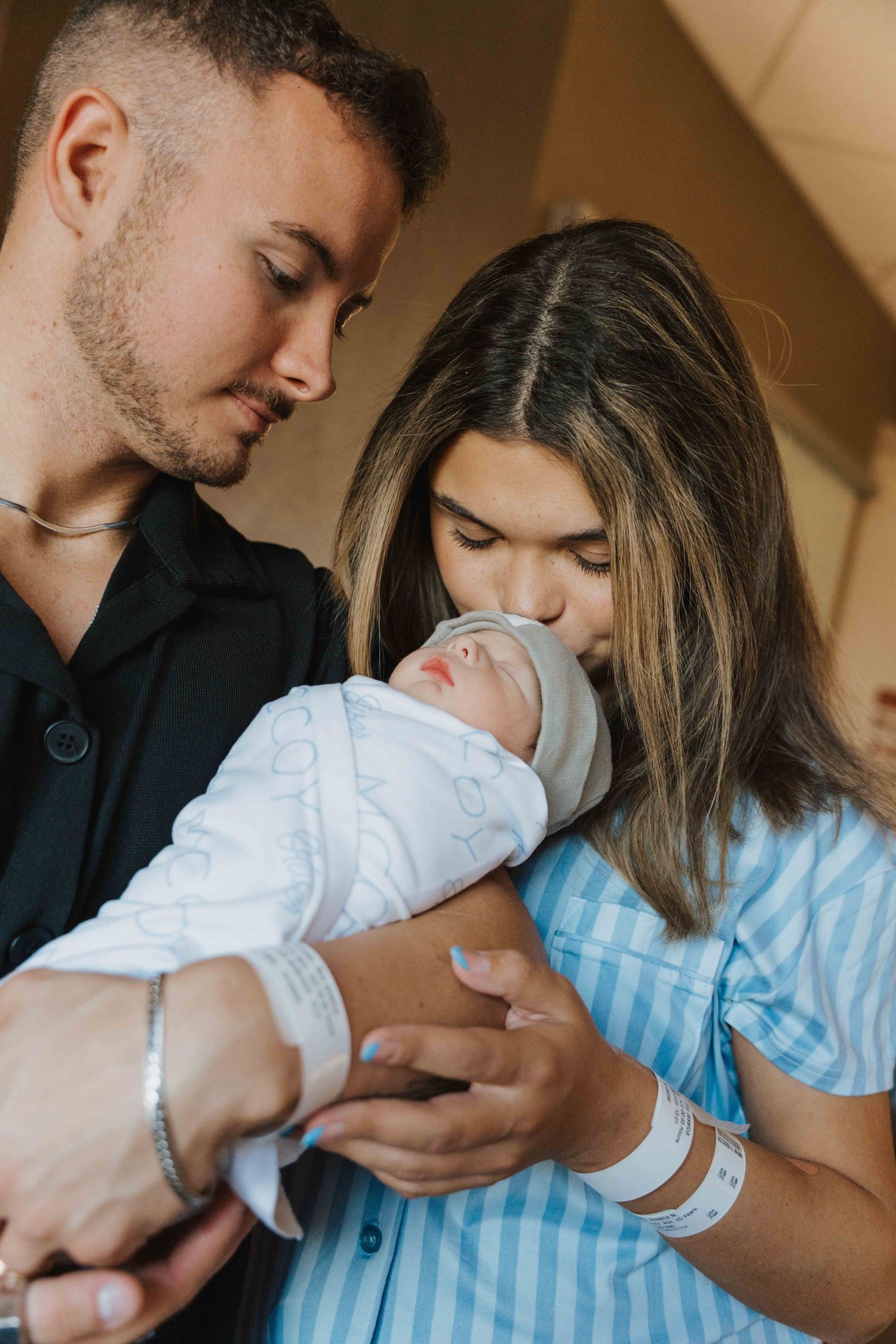 A couple holds and kisses their newborn baby in a series of tender moments captured in warm indoor lighting.