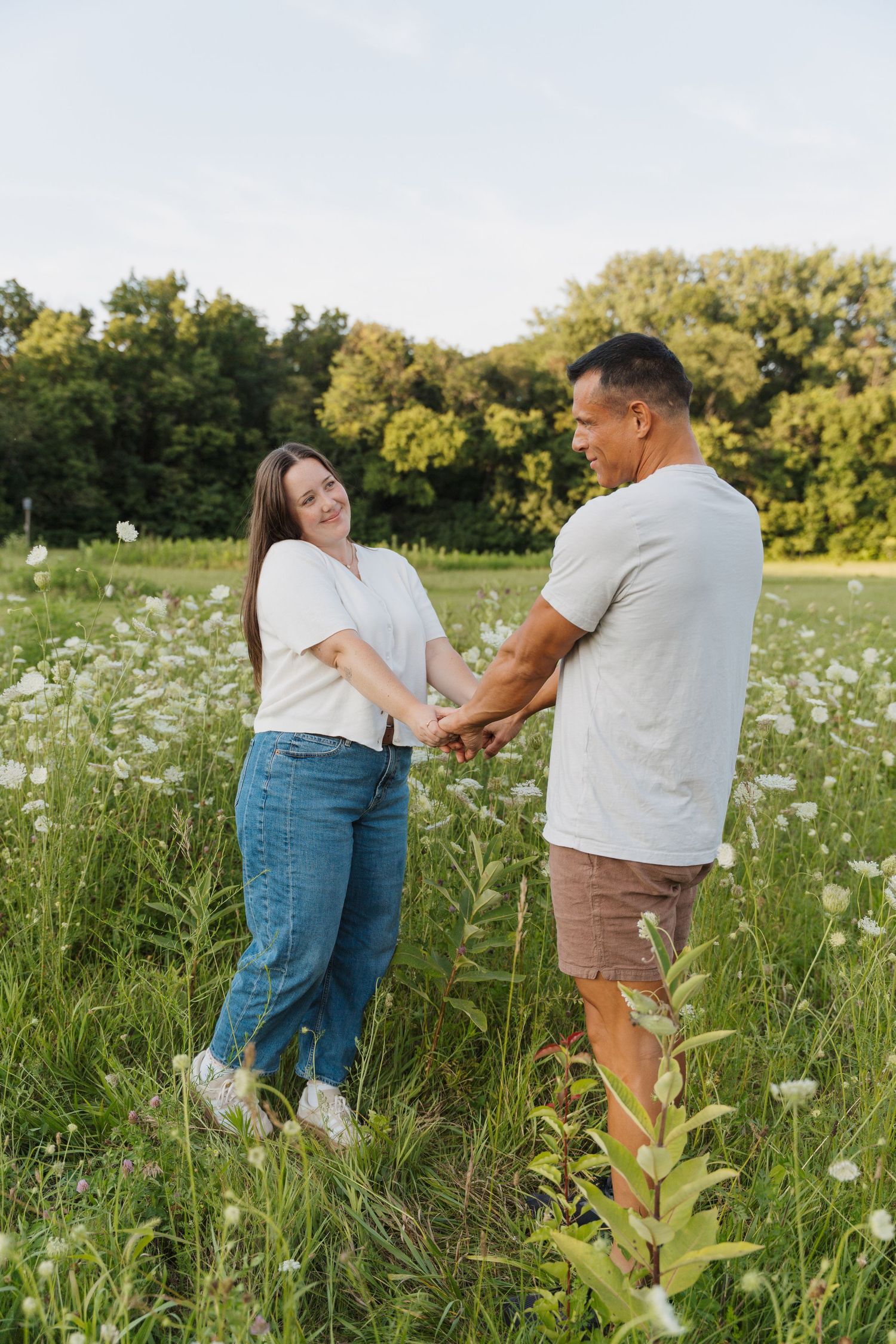 Happy couple holding hands in a blooming wildflower meadow with lush green trees in the background.