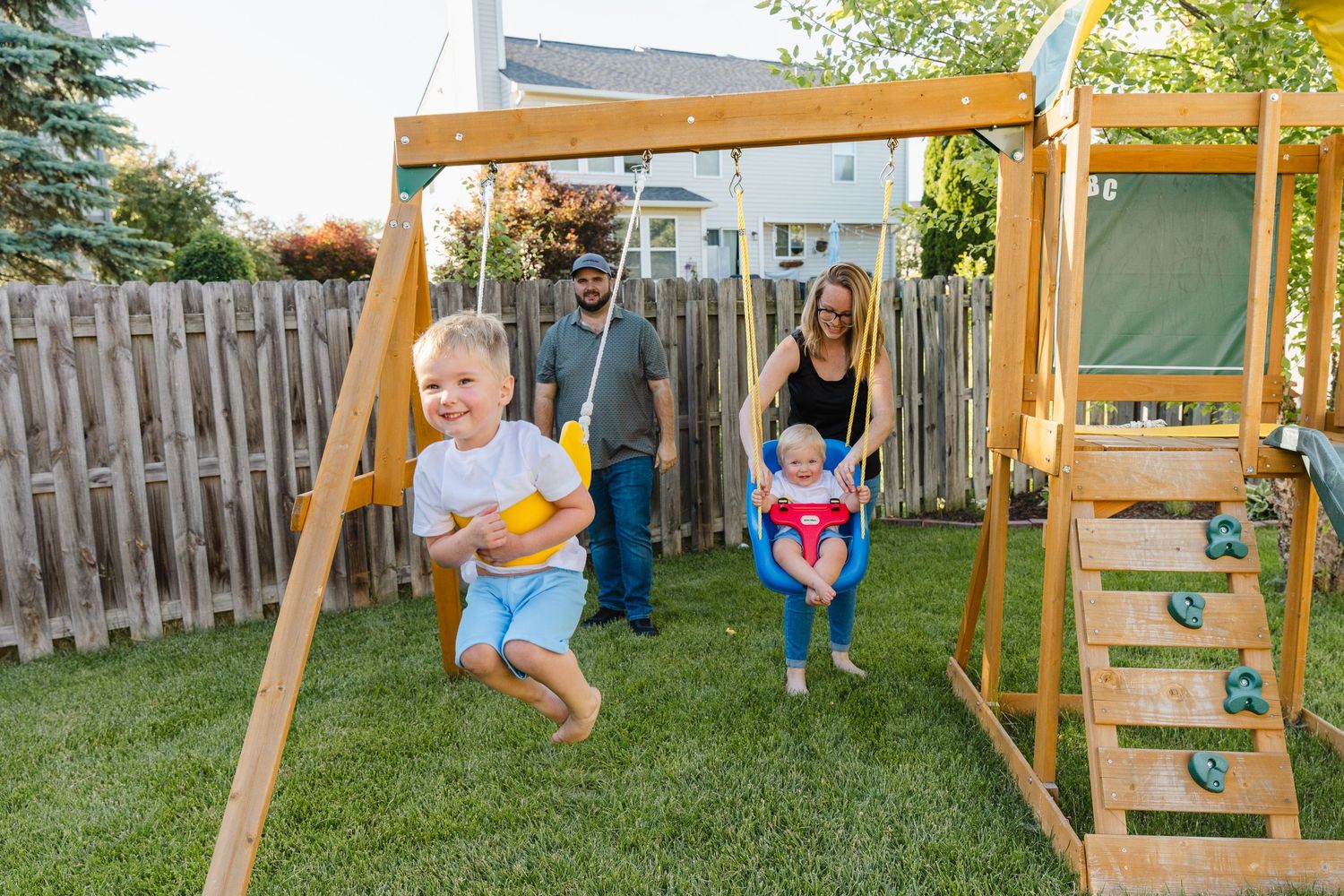 Family enjoying backyard playtime on wooden swing set, parents pushing young children on swings in sunny yard.