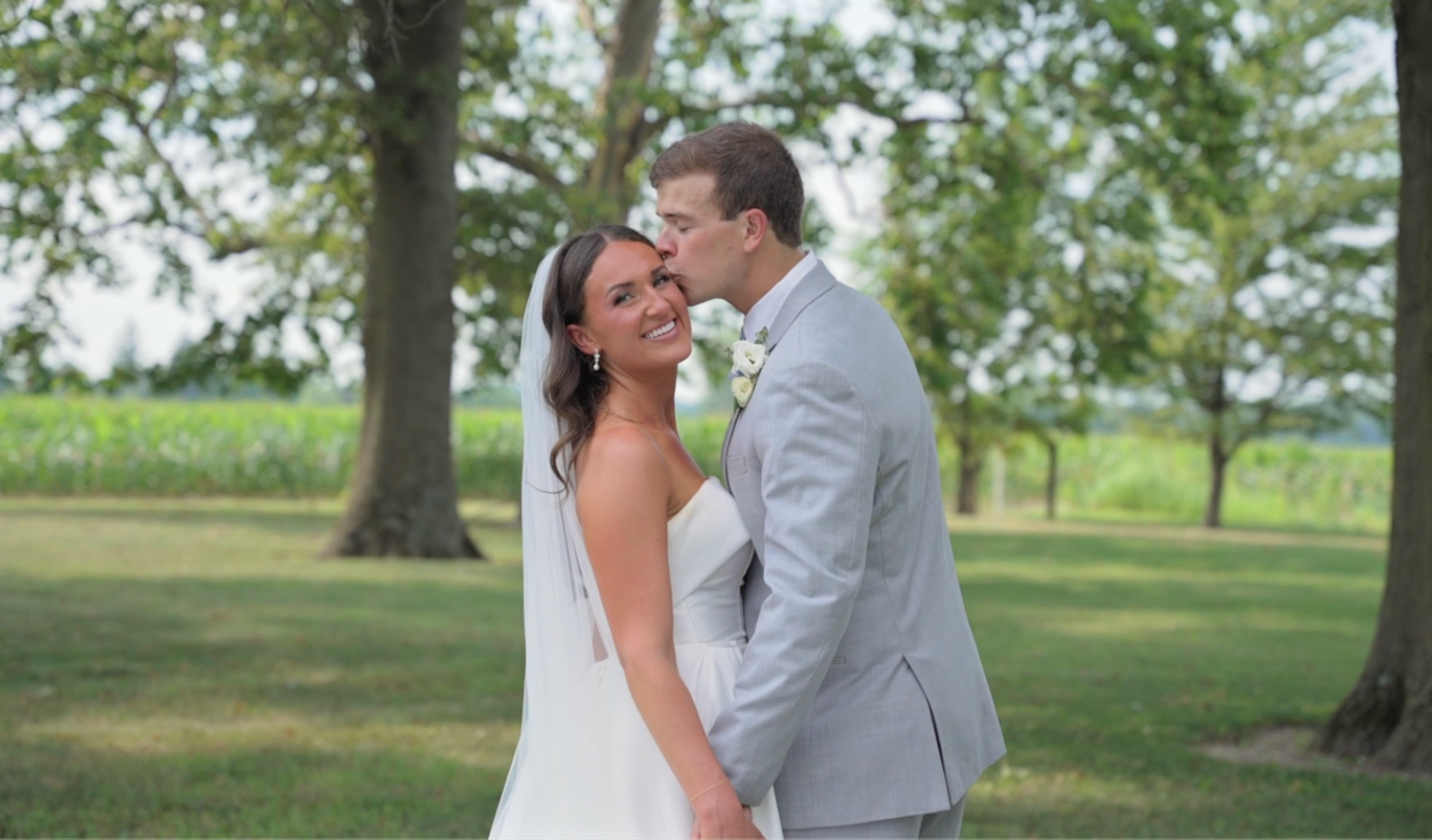 A couple in wedding attire share a romantic moment together on a grassy lawn with trees in the background.