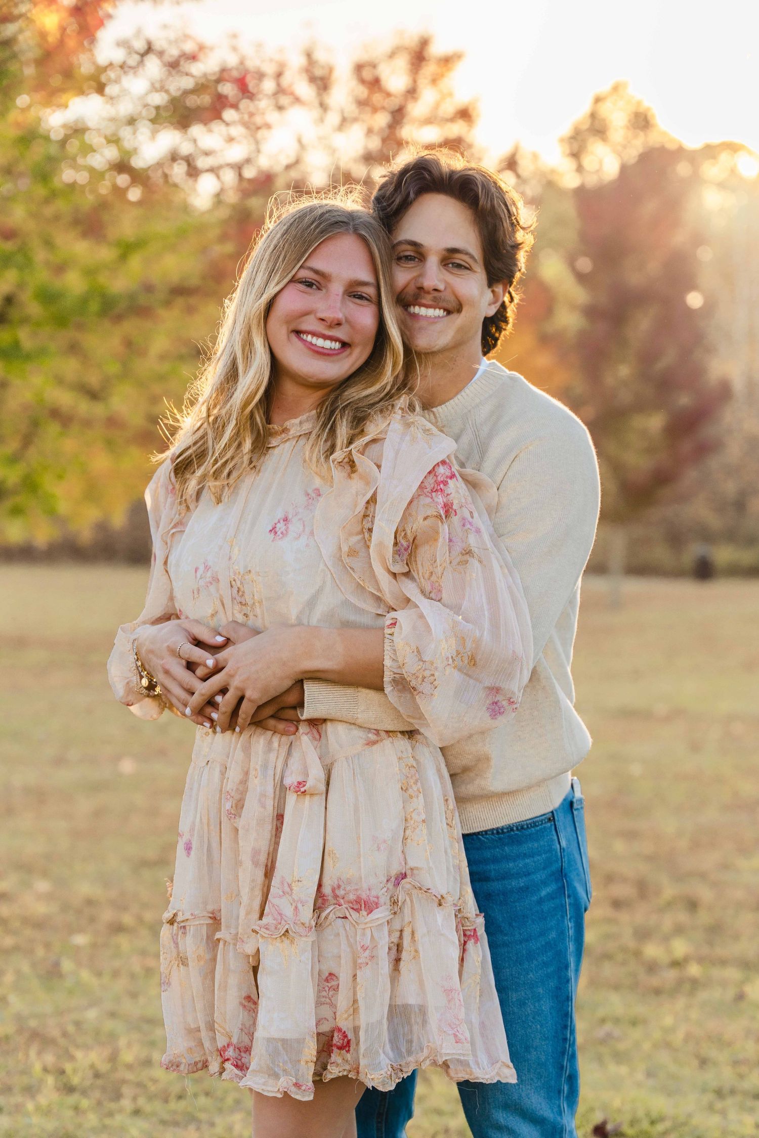 A couple embraces in a sunlit field during golden hour wearing floral dress and casual attire.