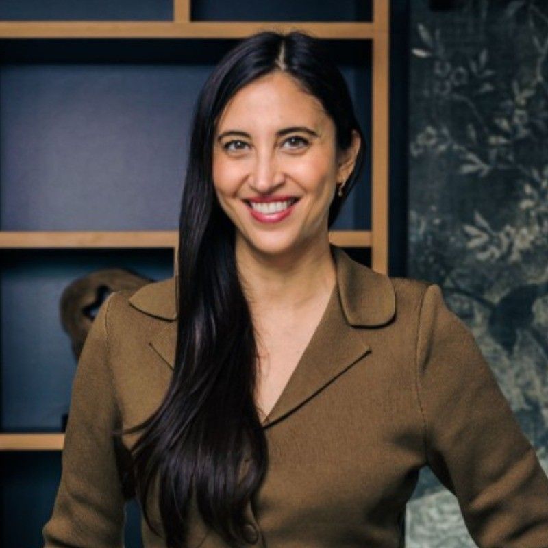 Professional woman with long dark hair smiling confidently in a brown blazer against a modern office backdrop.