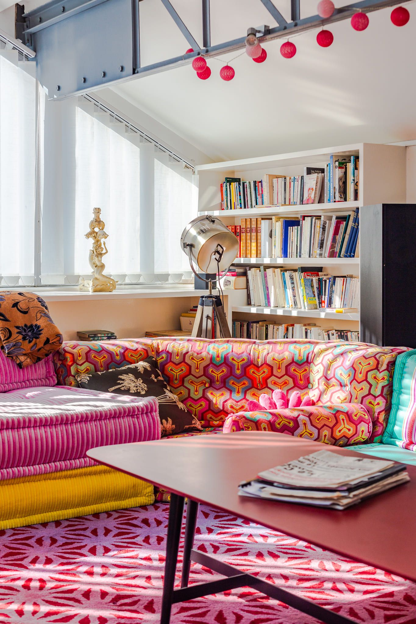 Colorful living room with vibrant patterned sofa, pink rug, bookshelf, and festive string lights.