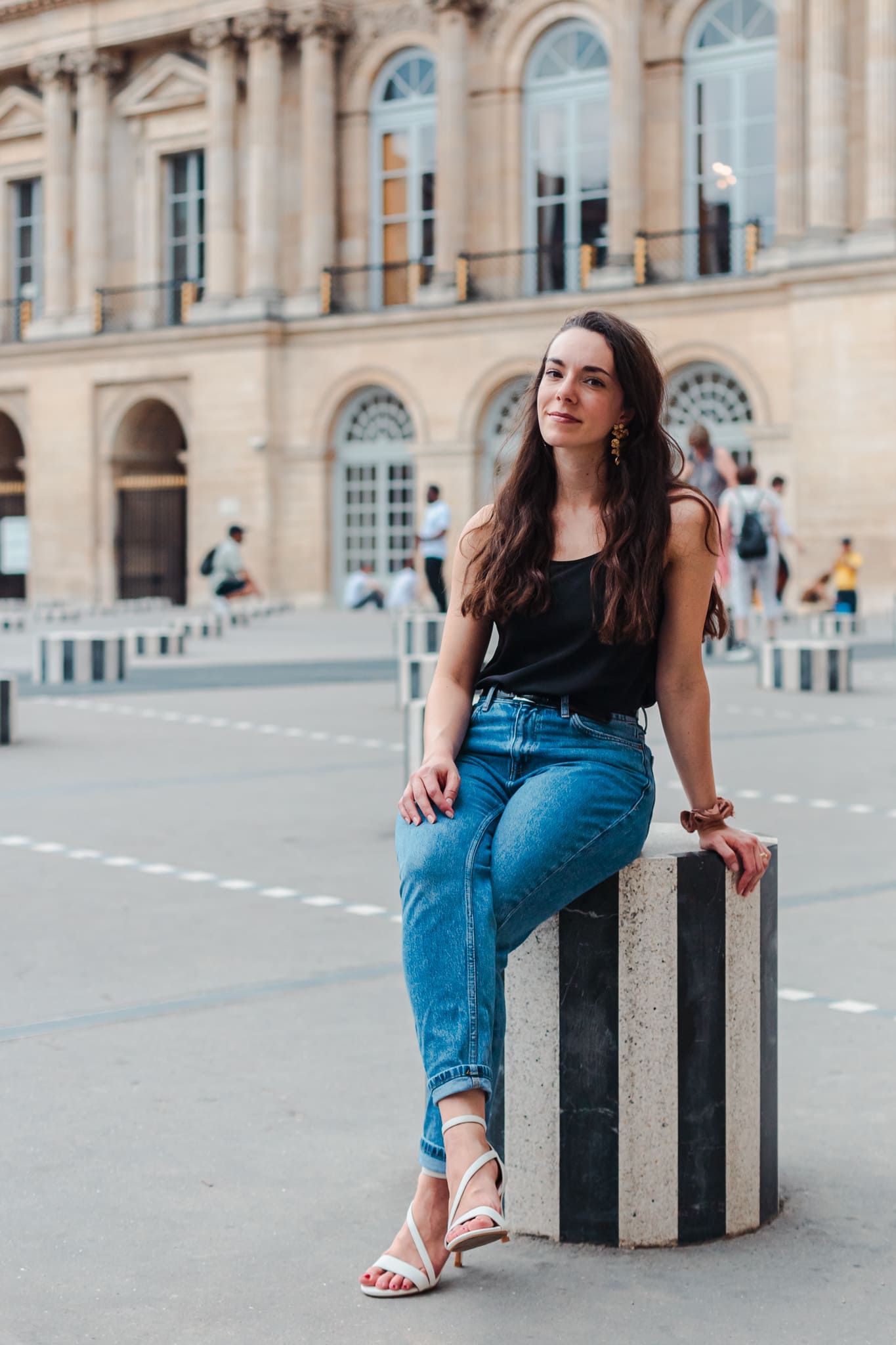 Young woman in black tank top and blue jeans sitting on striped bollard in front of classic European building.