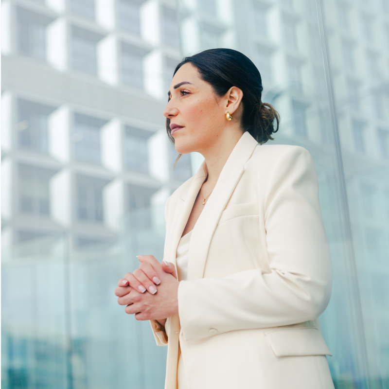 Professional woman in white blazer standing outdoors near modern glass building with hands clasped.