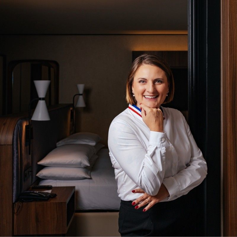 Smiling woman in white chef coat with French tricolor ribbon standing in elegant hotel room doorway.
