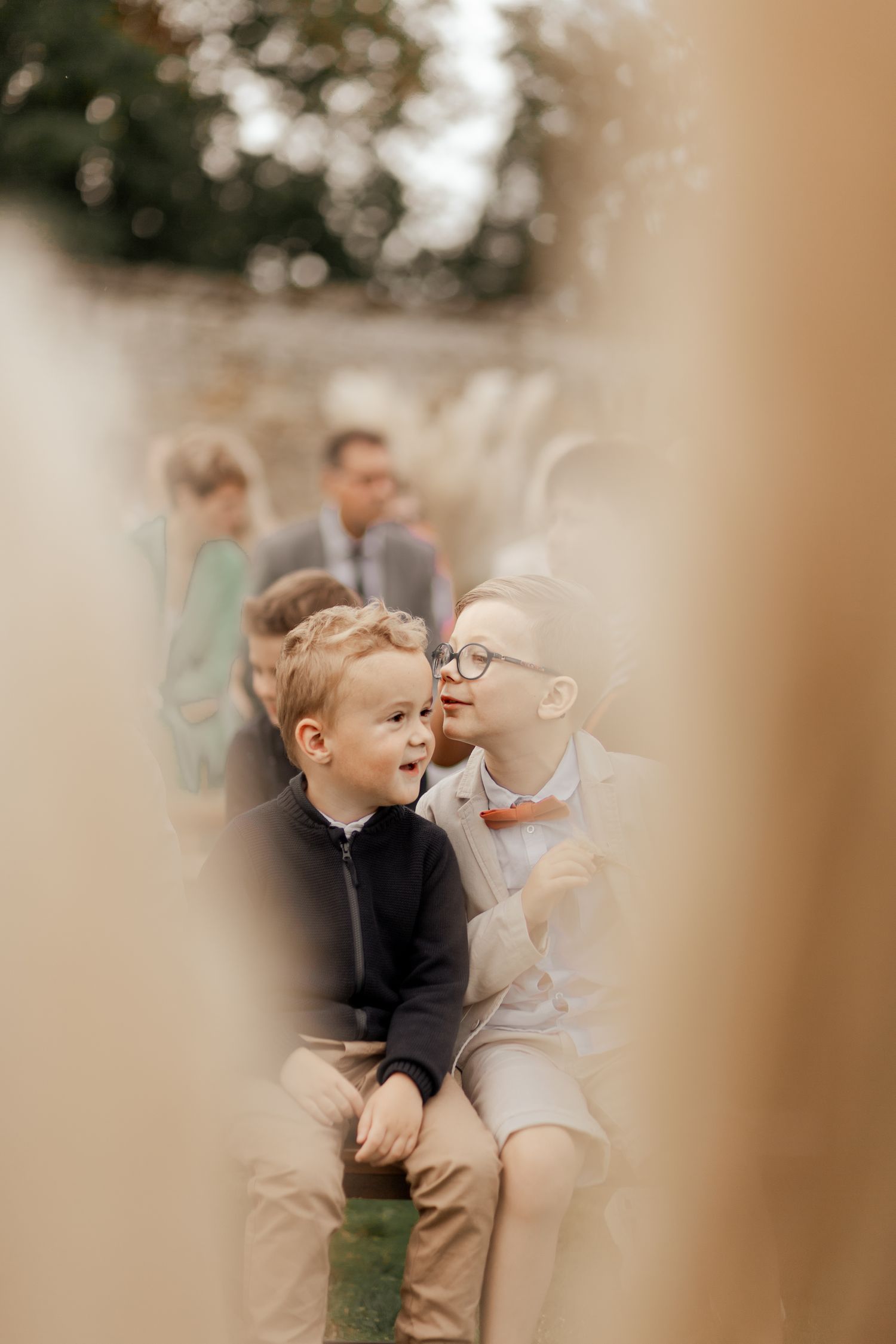 Two young boys in formal attire sit together at an outdoor wedding ceremony, whispering and smiling.