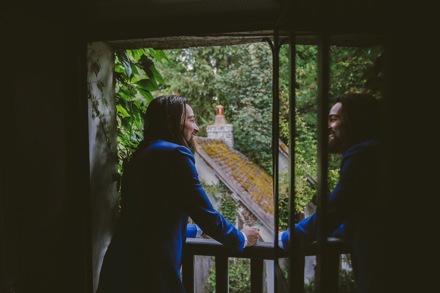 Couple in blue jackets sharing a romantic moment on a rustic balcony surrounded by lush green trees.