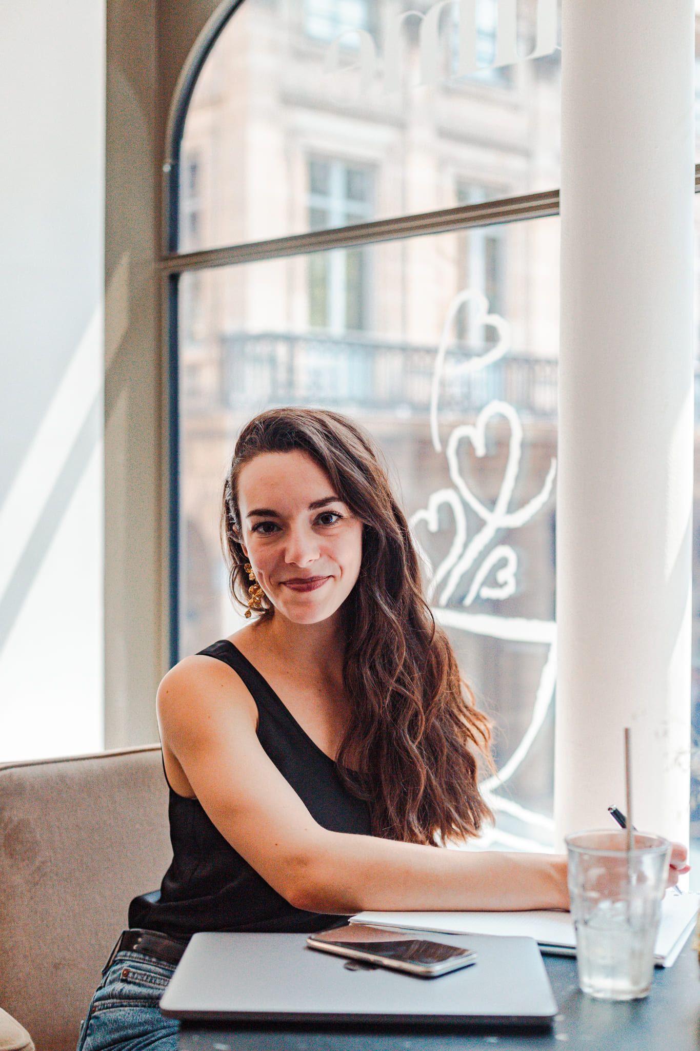 Young woman with long brown hair smiling in a cafe, sitting at a table with a laptop and glass of water.