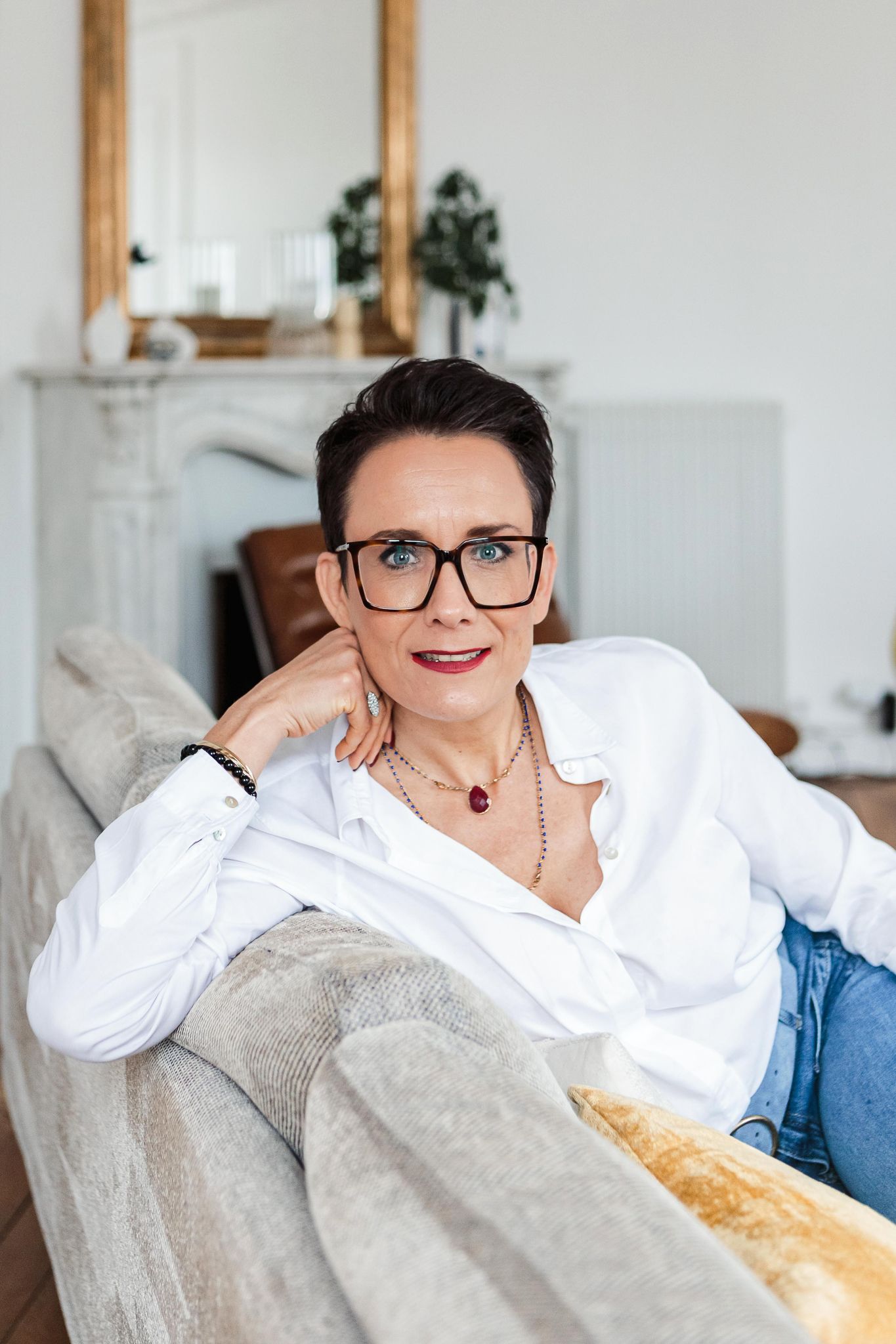 Stylish mature woman with short hair and glasses relaxes on sofa in bright Scandinavian living room.