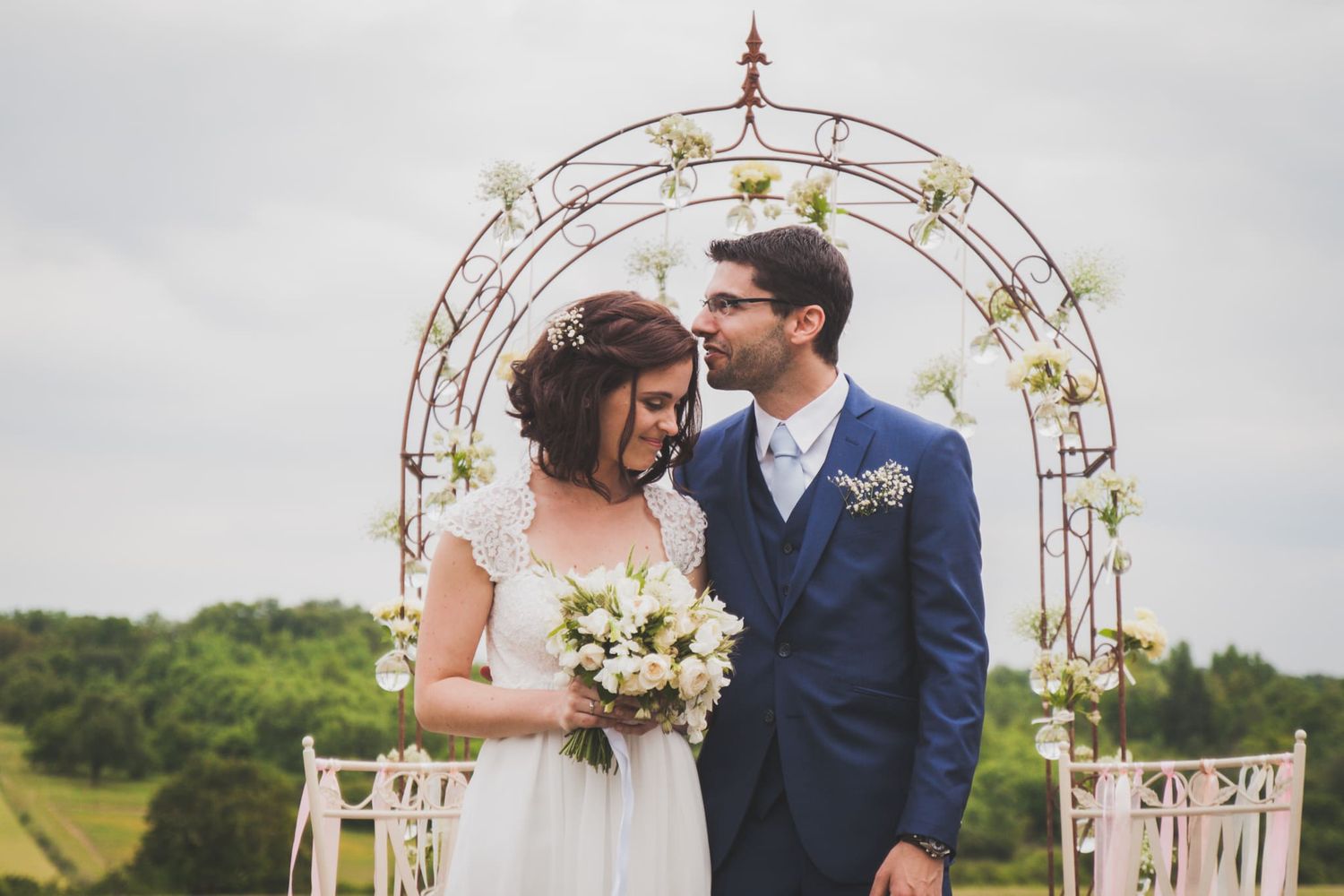 Bride and groom share a tender moment under a floral arch at an outdoor countryside wedding ceremony.