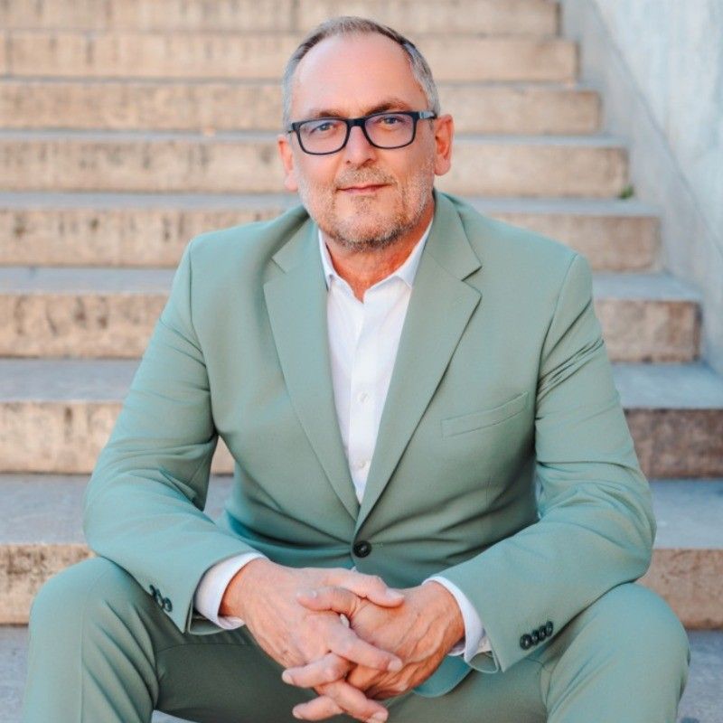 Confident middle-aged man in sage green suit sitting on stone steps with hands clasped.