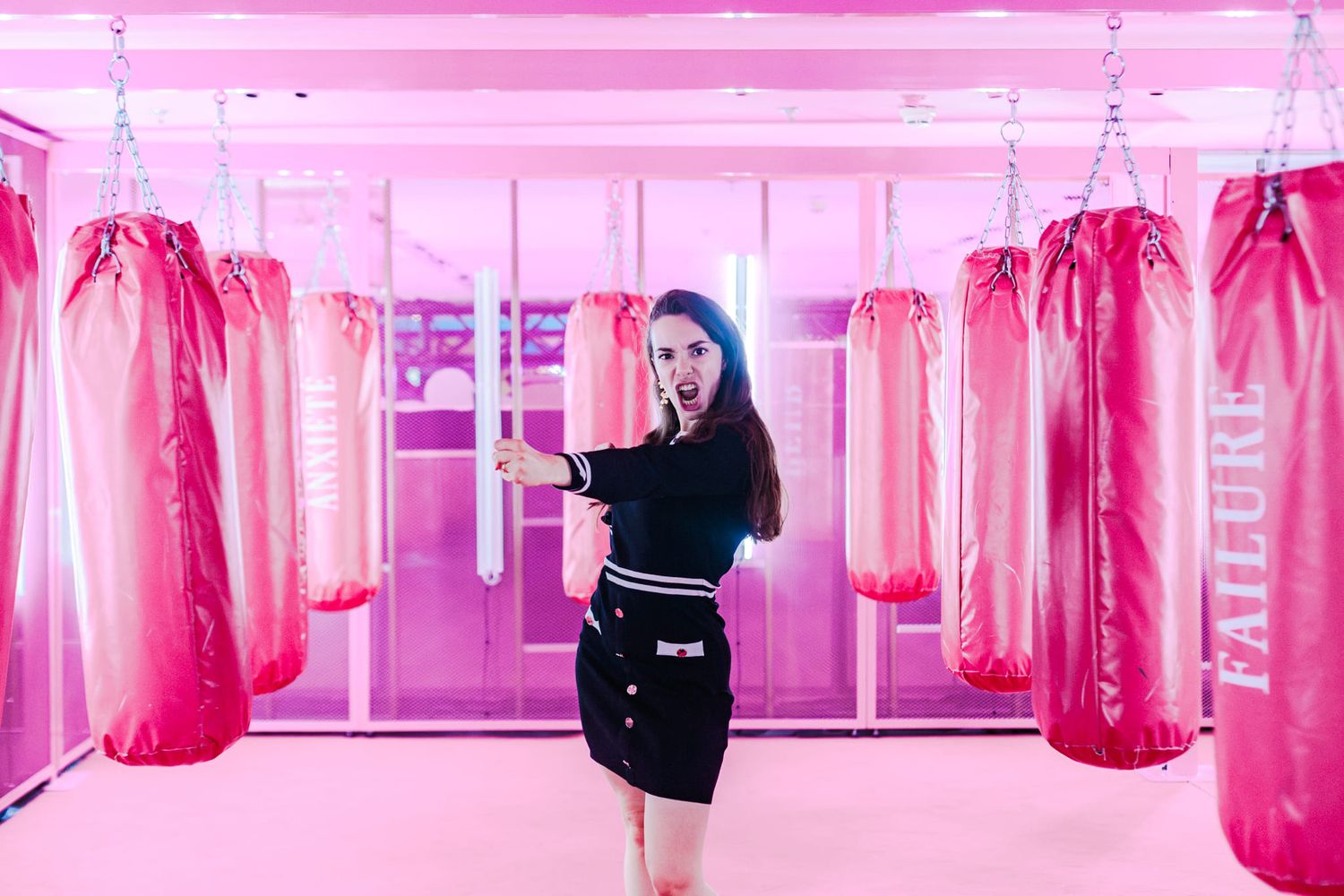 Woman in black outfit poses among pink punching bags in a vibrant pink boxing gym with neon lighting.