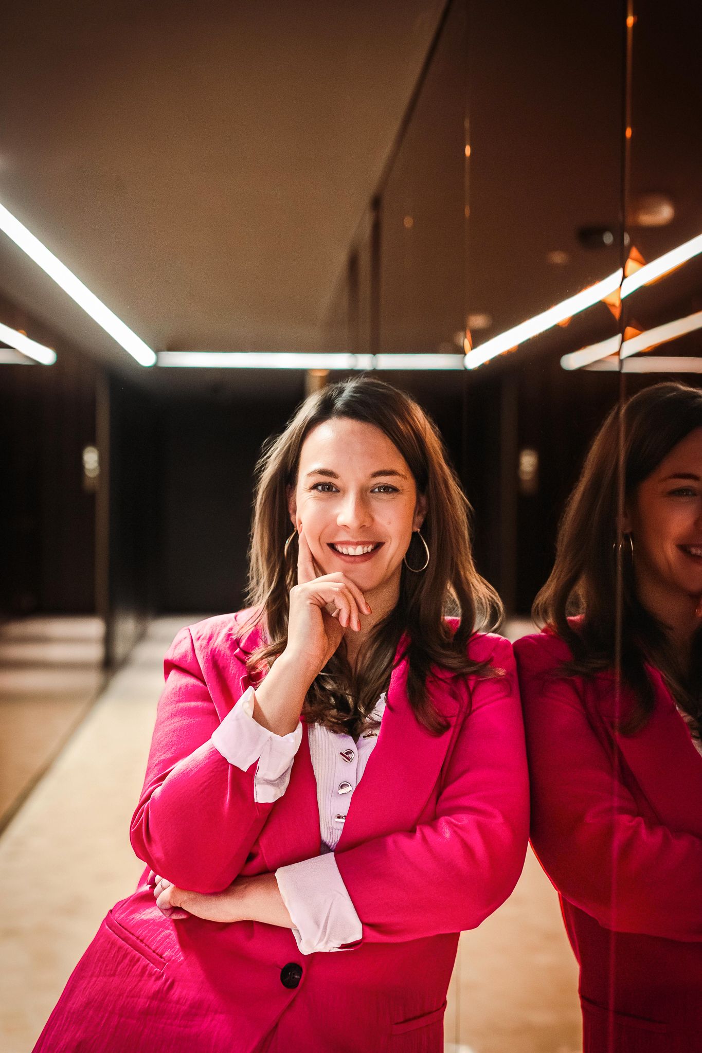 Smiling woman in a bright pink blazer leaning against a mirror in a modern interior with warm lighting.