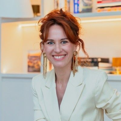Professional headshot of a smiling redhead woman in a white blazer in a bright modern interior.