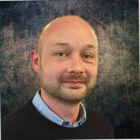 Bald man in black sweater with professional headshot against textured background.