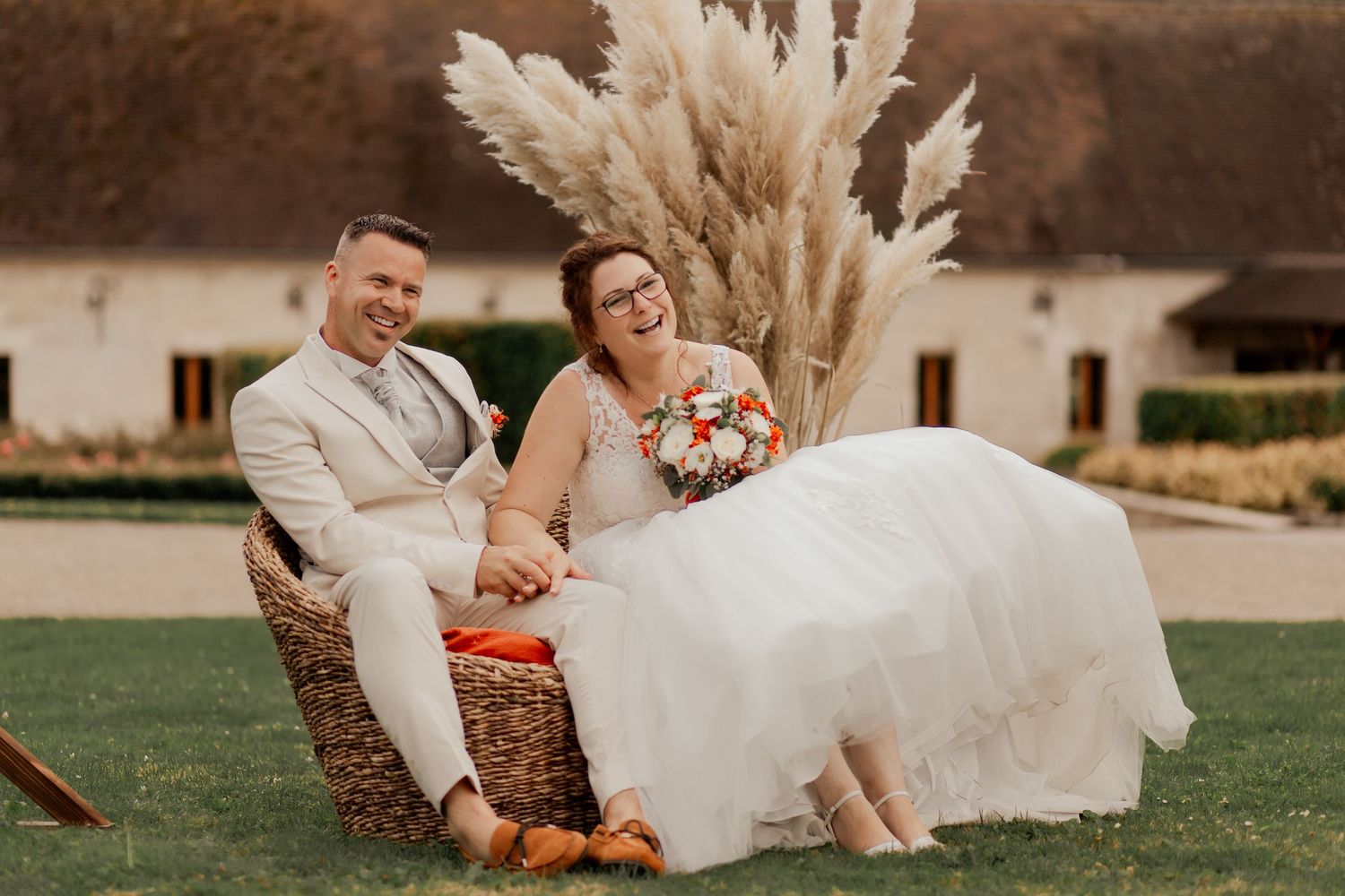 Happy bride and groom sitting in wicker chair with pampas grass backdrop on wedding day outdoors.