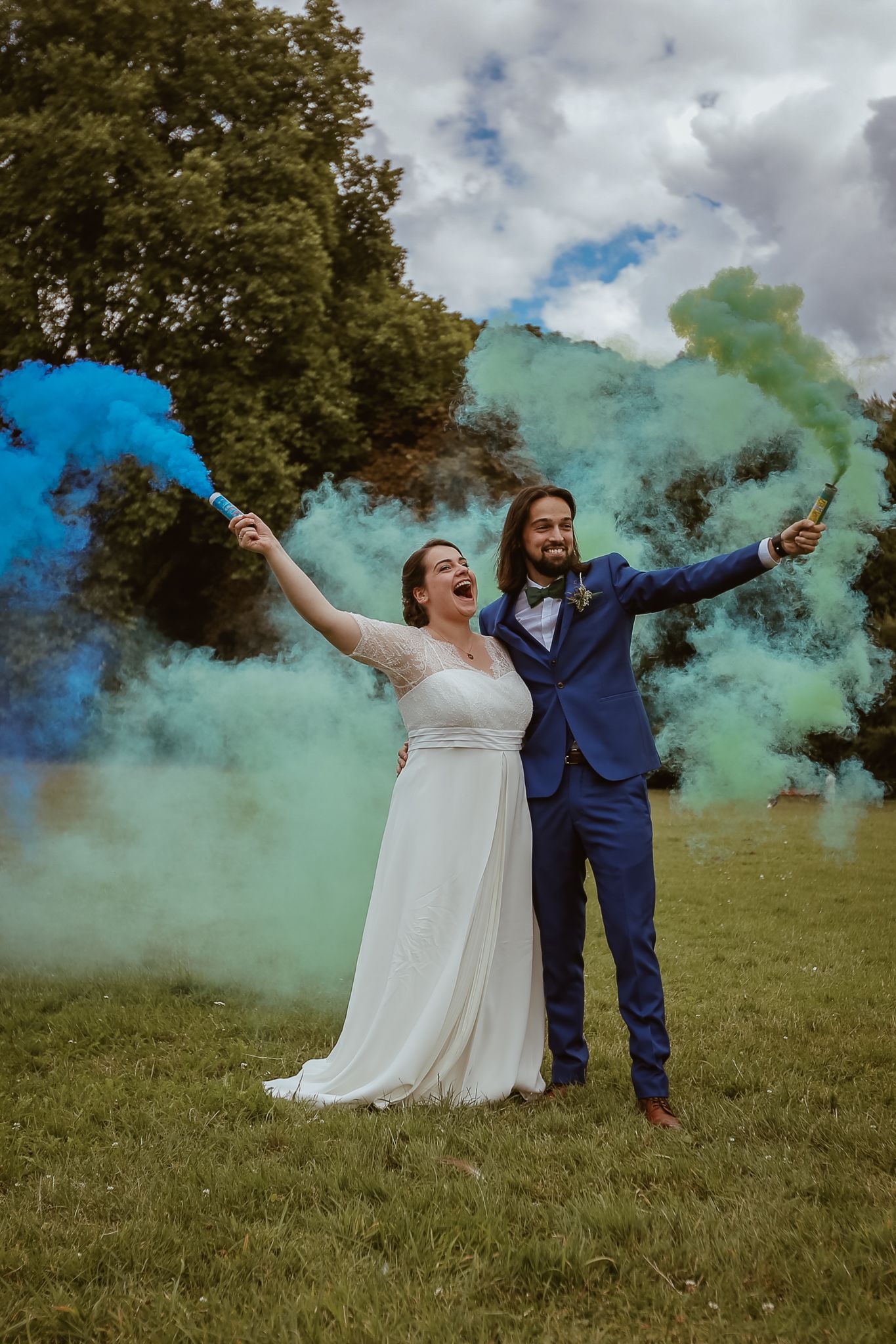 Bride and groom celebrate outdoors holding colorful blue and green smoke bombs in a lush green field on their wedding day.