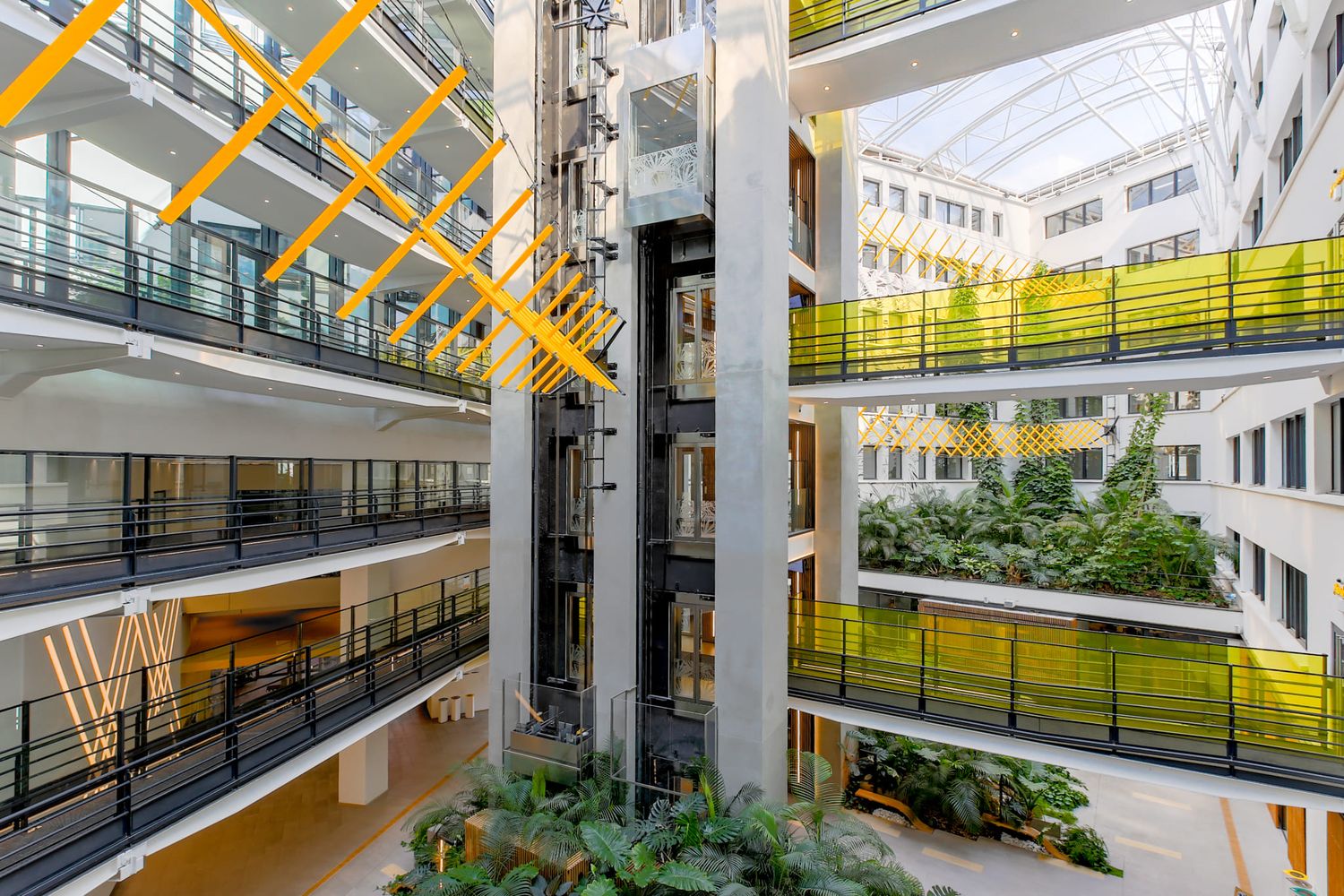 Modern office atrium with yellow structural beams, green plant walls, and multi-level balconies.