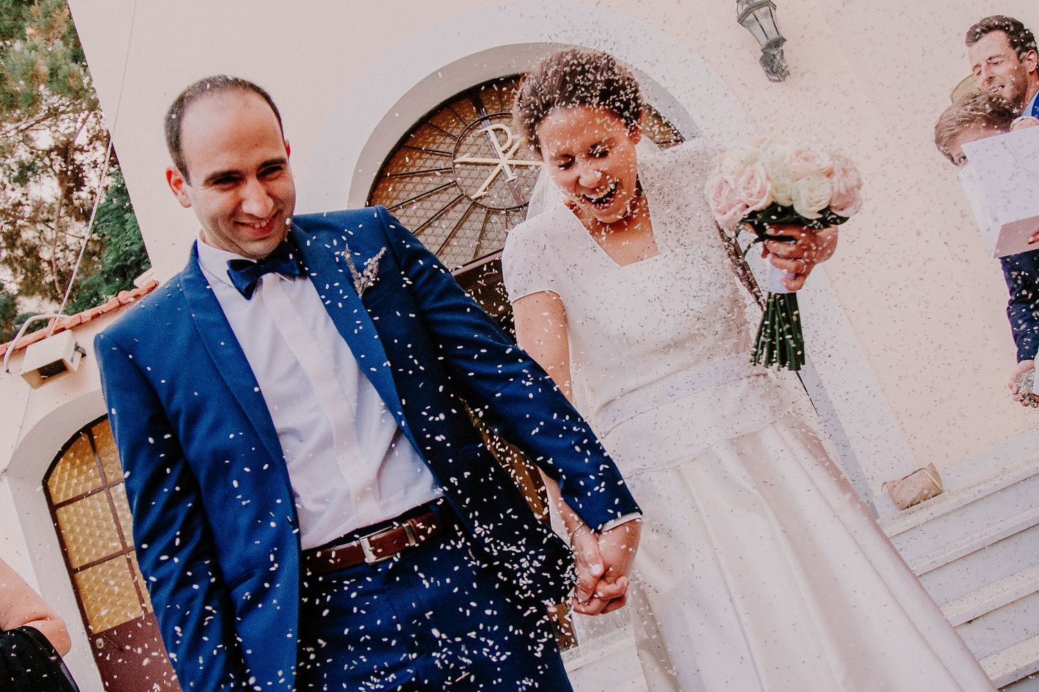 Joyful newlyweds exit church in confetti shower, groom in blue suit, bride in white gown holding bouquet.