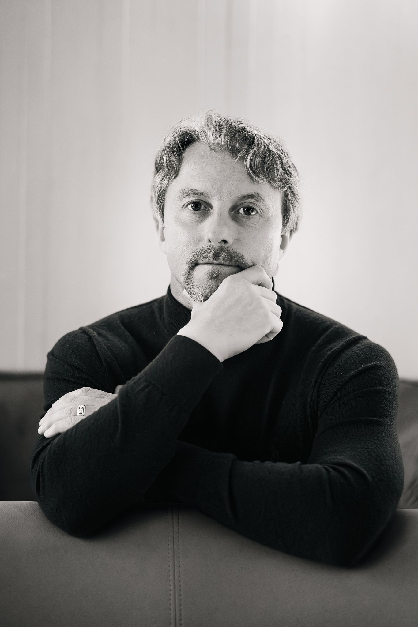 Black and white portrait of a man in black turtleneck seated on a leather sofa with chin resting on hand.