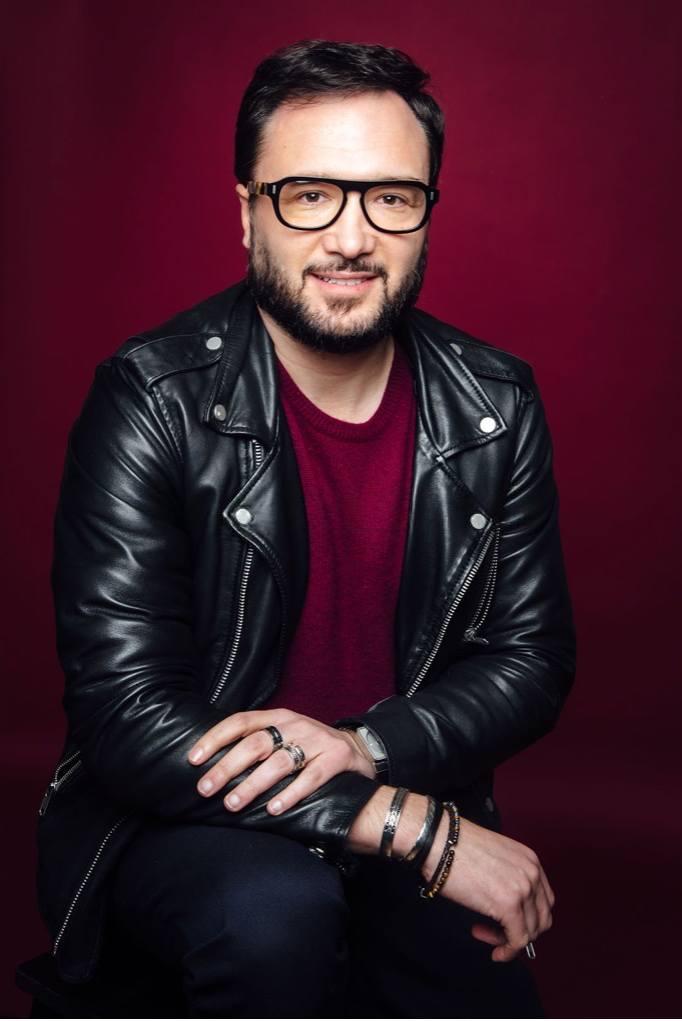 Confident man wearing black leather jacket, glasses, and bracelets posing against a rich burgundy studio backdrop.