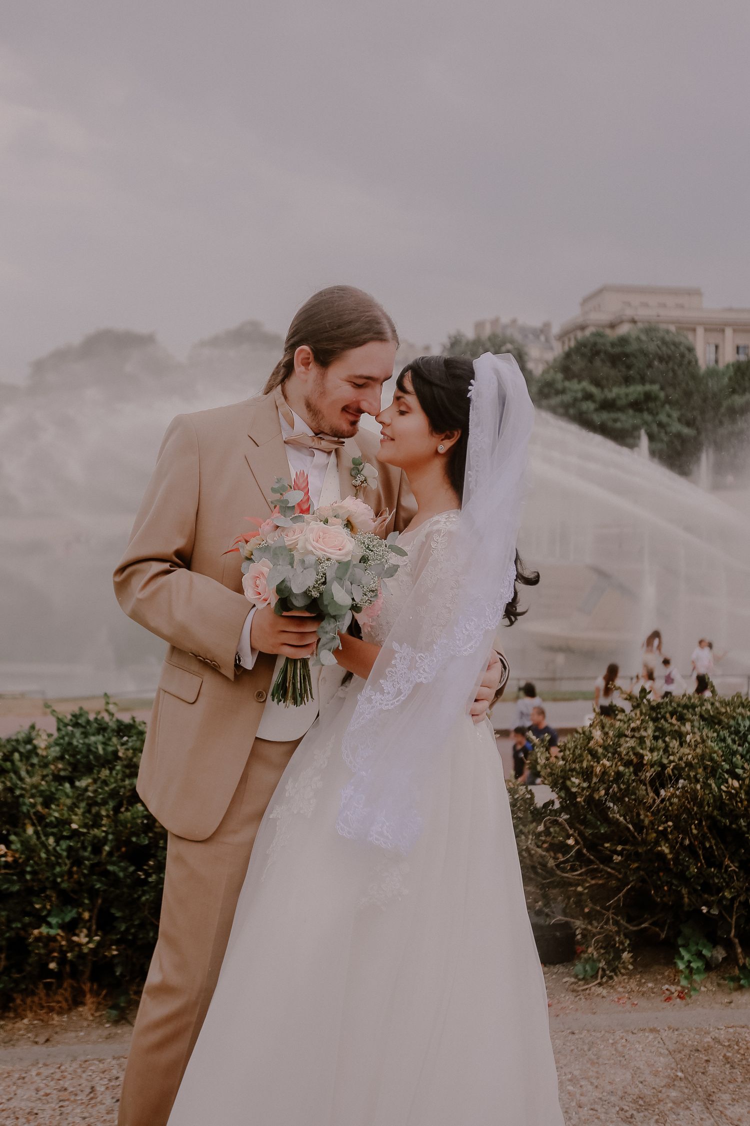 Bride and groom share a romantic moment, she in white lace gown with veil, he in tan suit, holding a bouquet outdoors.