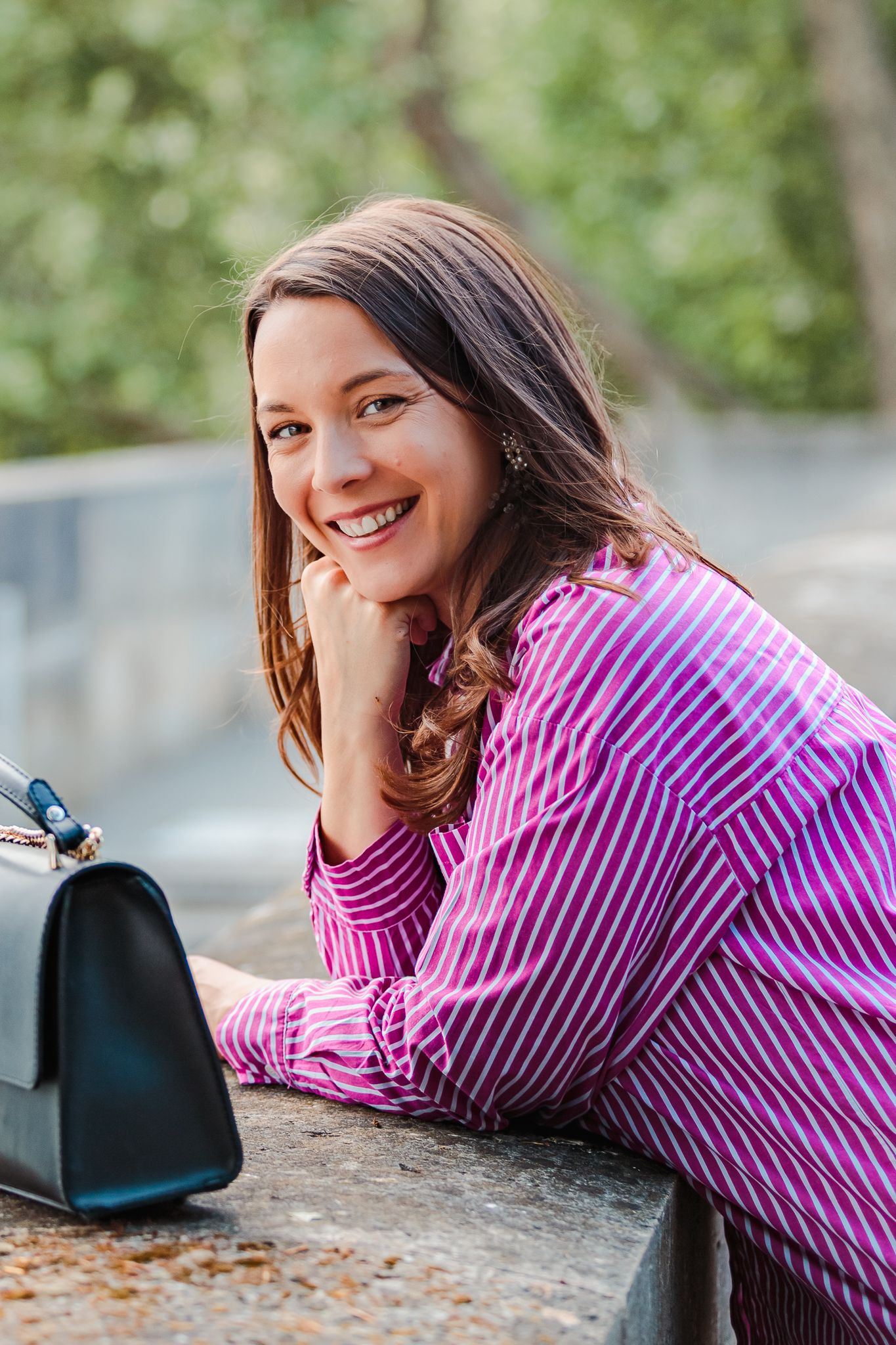 Smiling woman in pink striped shirt leaning on stone ledge outdoors with black handbag beside her.